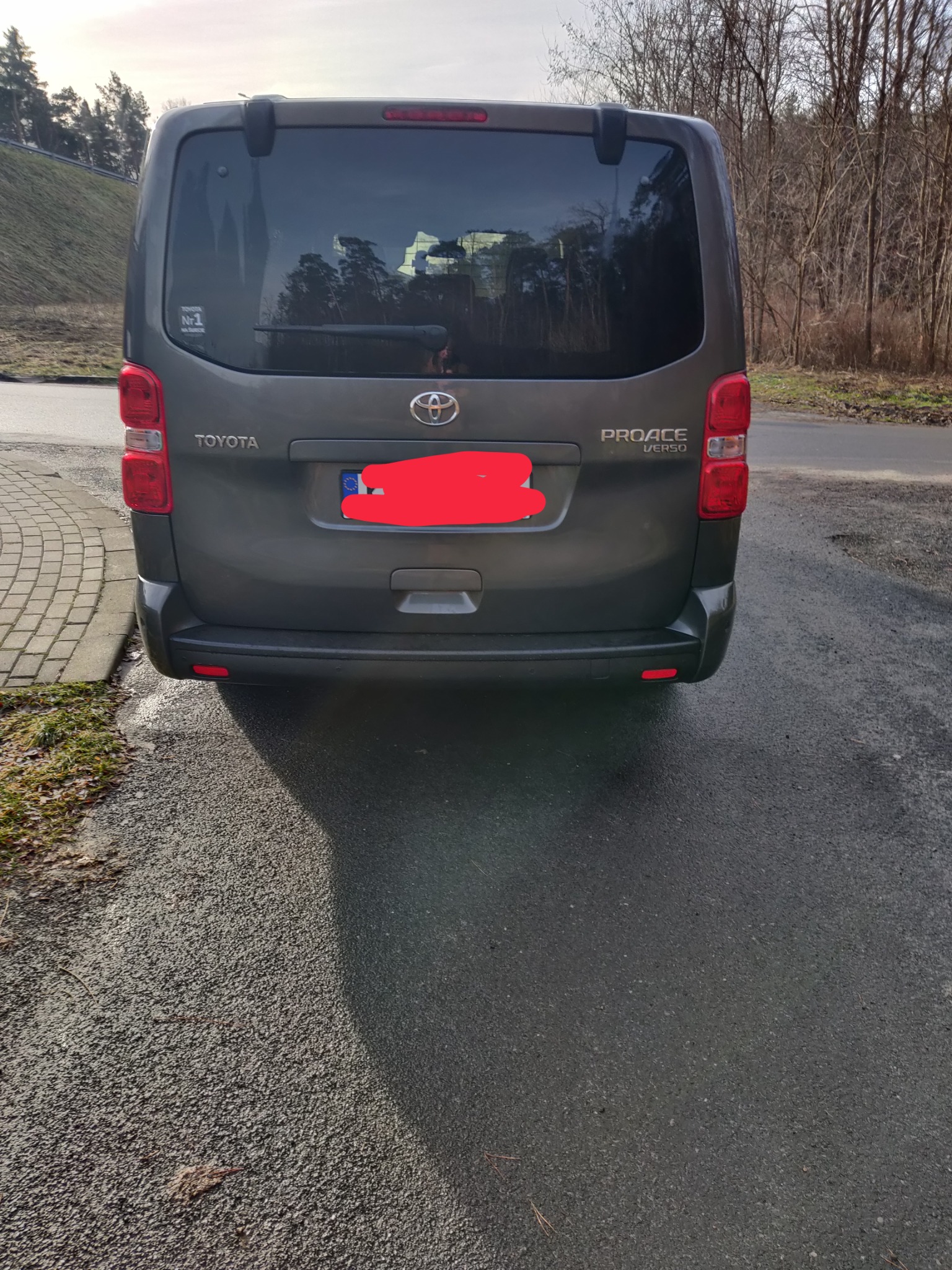 Tył szarego samochodu Toyota Proace Verso z widoczną naklejką 'Nr 1 na wakacje' na szybie, zaparkowanego na asfalcie.