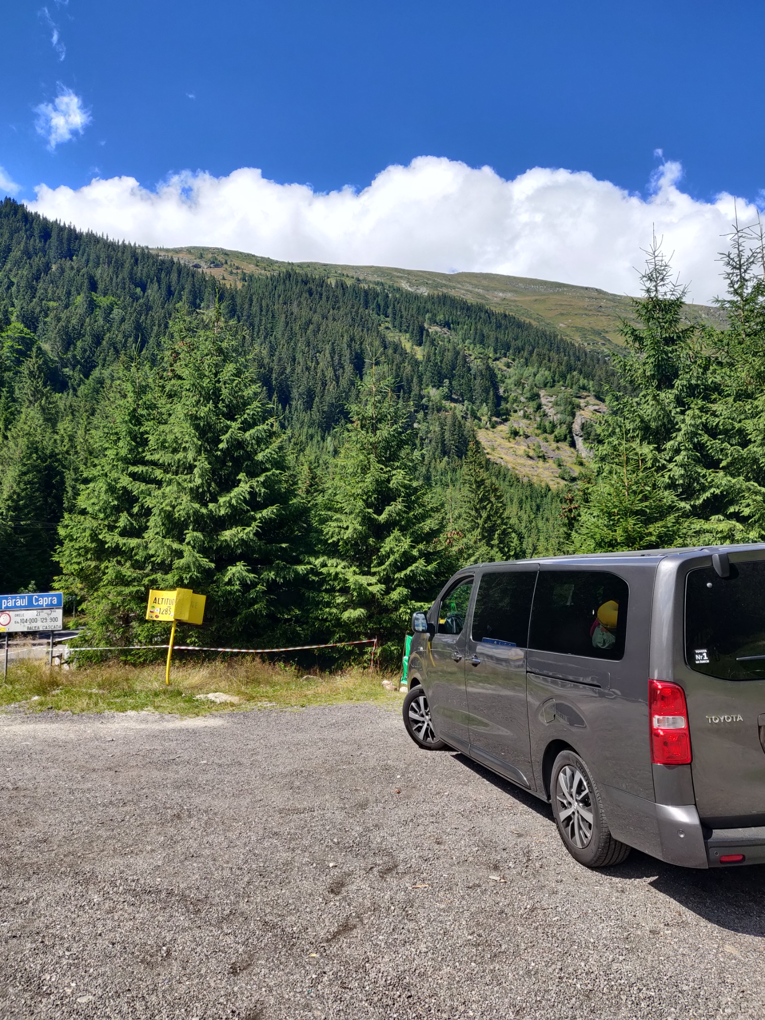 Szary van Toyota zaparkowany na żwirowym parkingu z widokiem na zalesione góry i tablicę informacyjną o wysokości nad poziomem morza w tle.