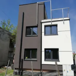 Dwukondygnacyjny budynek z elewacją w kolorze białym i grafitowym, widoczne okna z ciemnymi ramami, rura spustowa, niezakończone bariery na dachu i teren wokół budynku w trakcie prac.