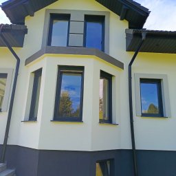 KLIM-BUD USŁUGI REMONTOWO BUDOWLANE Krzysztof Klimek - Elewacja domu jednorodzinnego po remoncie: jasna fasada z ciemną podmurówką i stolarką okienną. Widoczne rynny i fragment schodów wejściowych. Błękitne niebo odbija się w szybach.