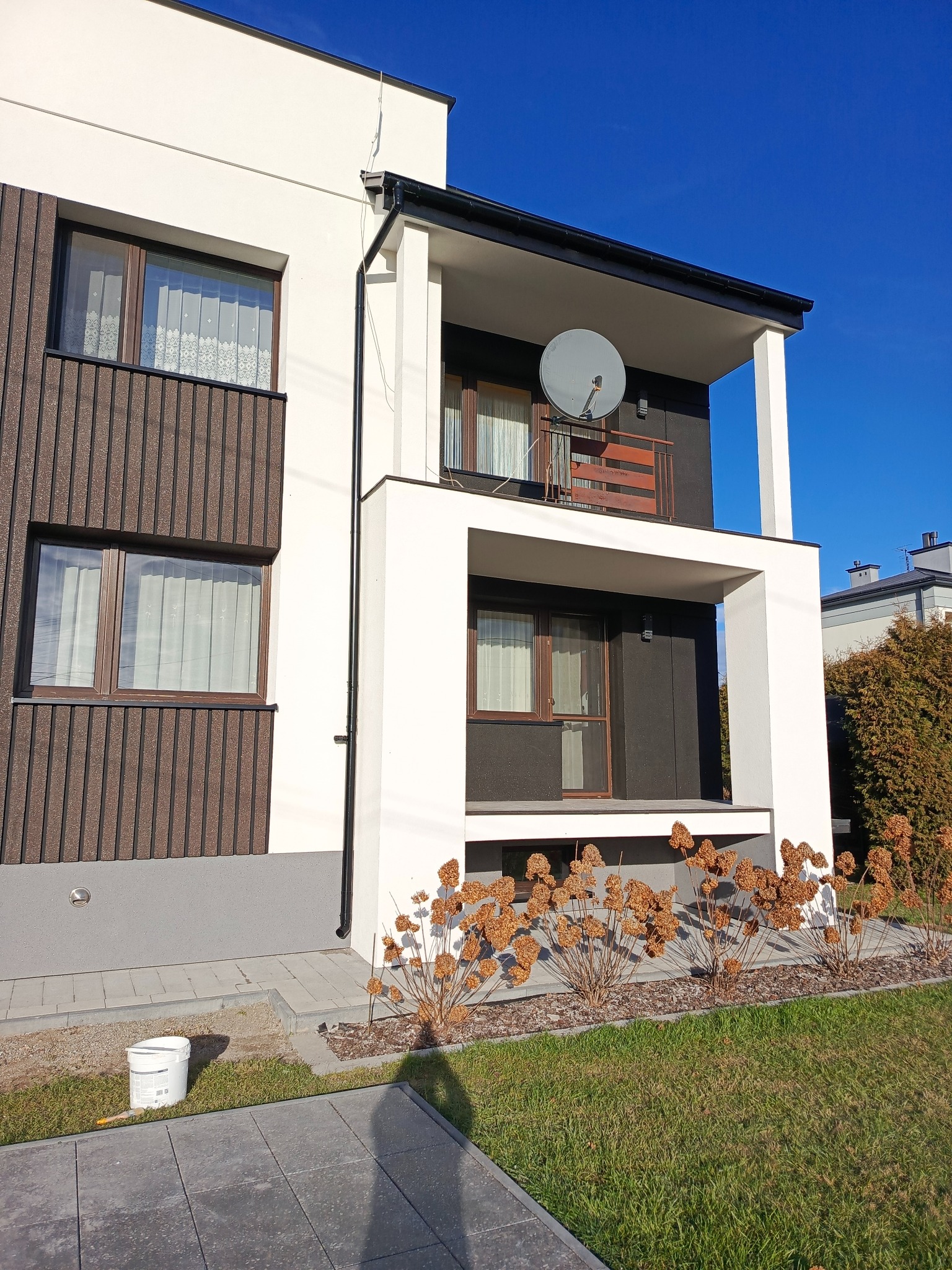 Dwukondygnacyjny dom z balkonem, elewacją w bieli i brązie, oraz anteną satelitarną. Widoczne detale wykończenia i otoczenie budynku w słoneczny dzień.