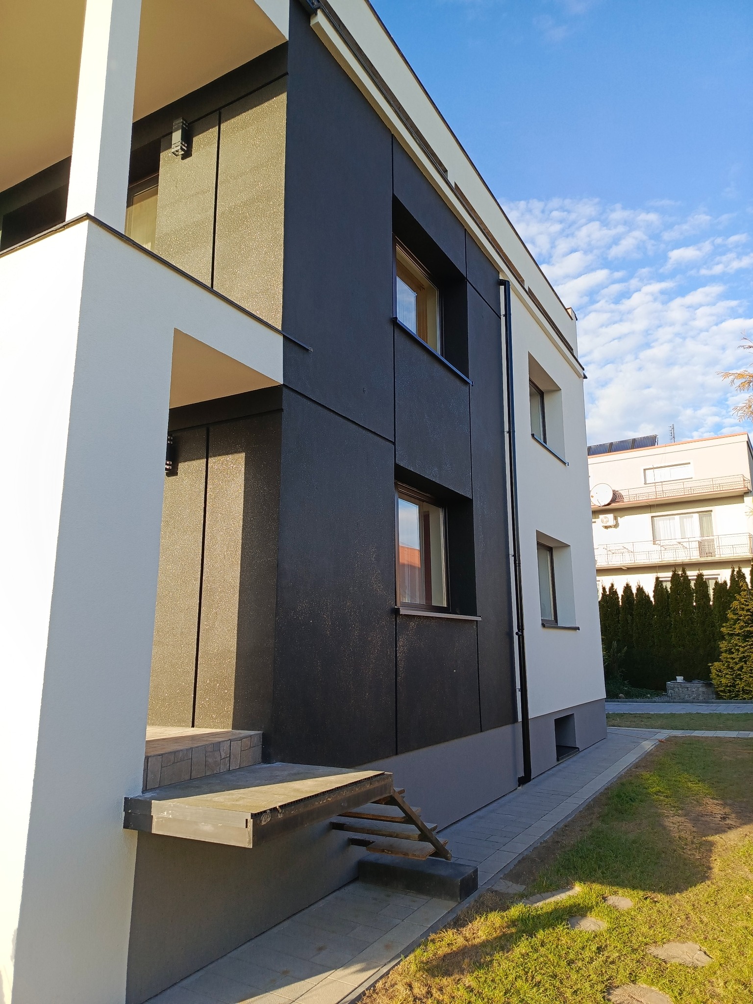 Nowoczesny budynek z elewacją w odcieniach bieli i grafitu, z widocznymi oknami i fragmentem balkonu. Ujęcie z dołu, podkreślające geometrię budynku na tle błękitnego nieba.