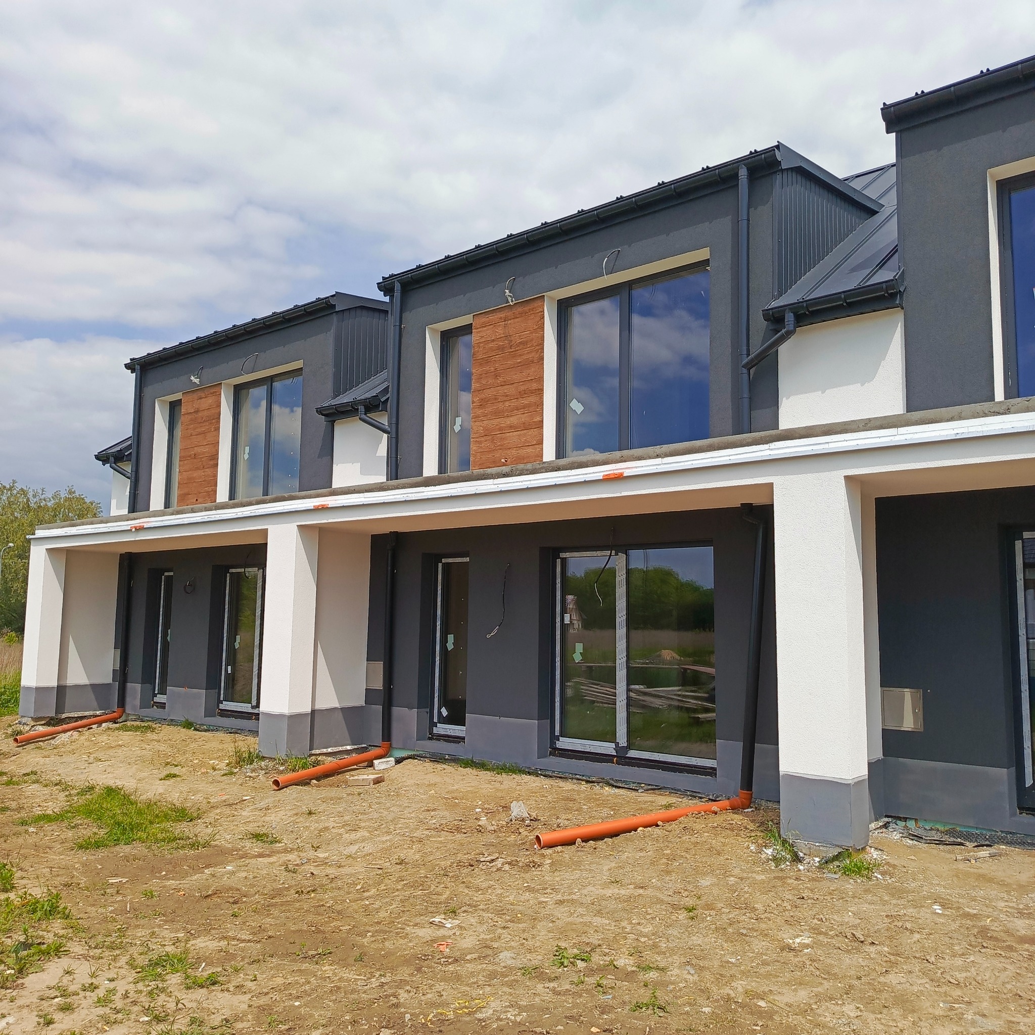 Nowoczesny szeregowiec z szarą elewacją, drewnianymi akcentami i dużymi oknami. Widoczne rury odprowadzające wodę. Nierówny teren przed budynkiem.