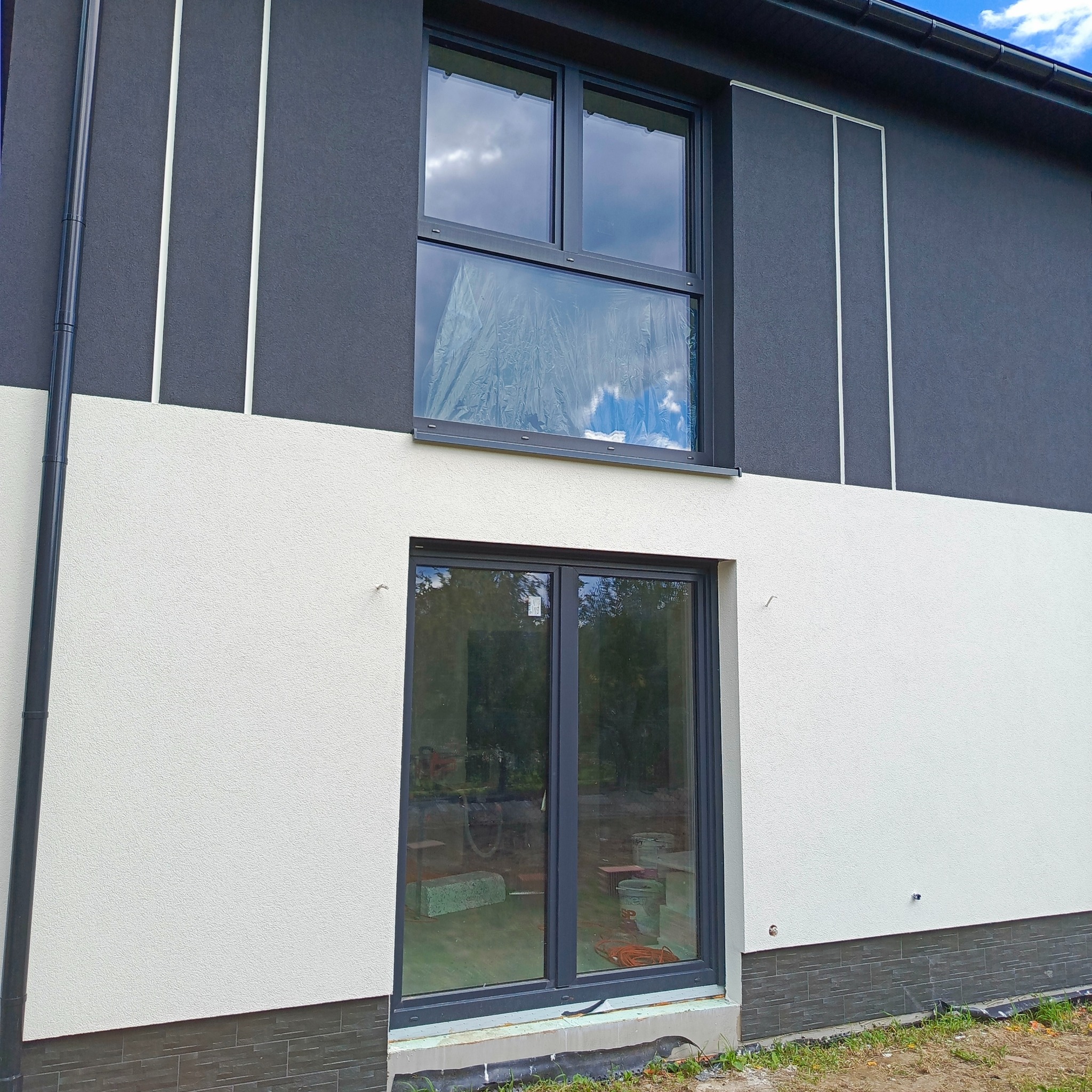 Dwukolorowa elewacja domu z szarymi oknami i dekoracyjnymi, pionowymi paskami. Okno na górze zabezpieczone folią. Widoczne detale wykończenia i fragment dachu.