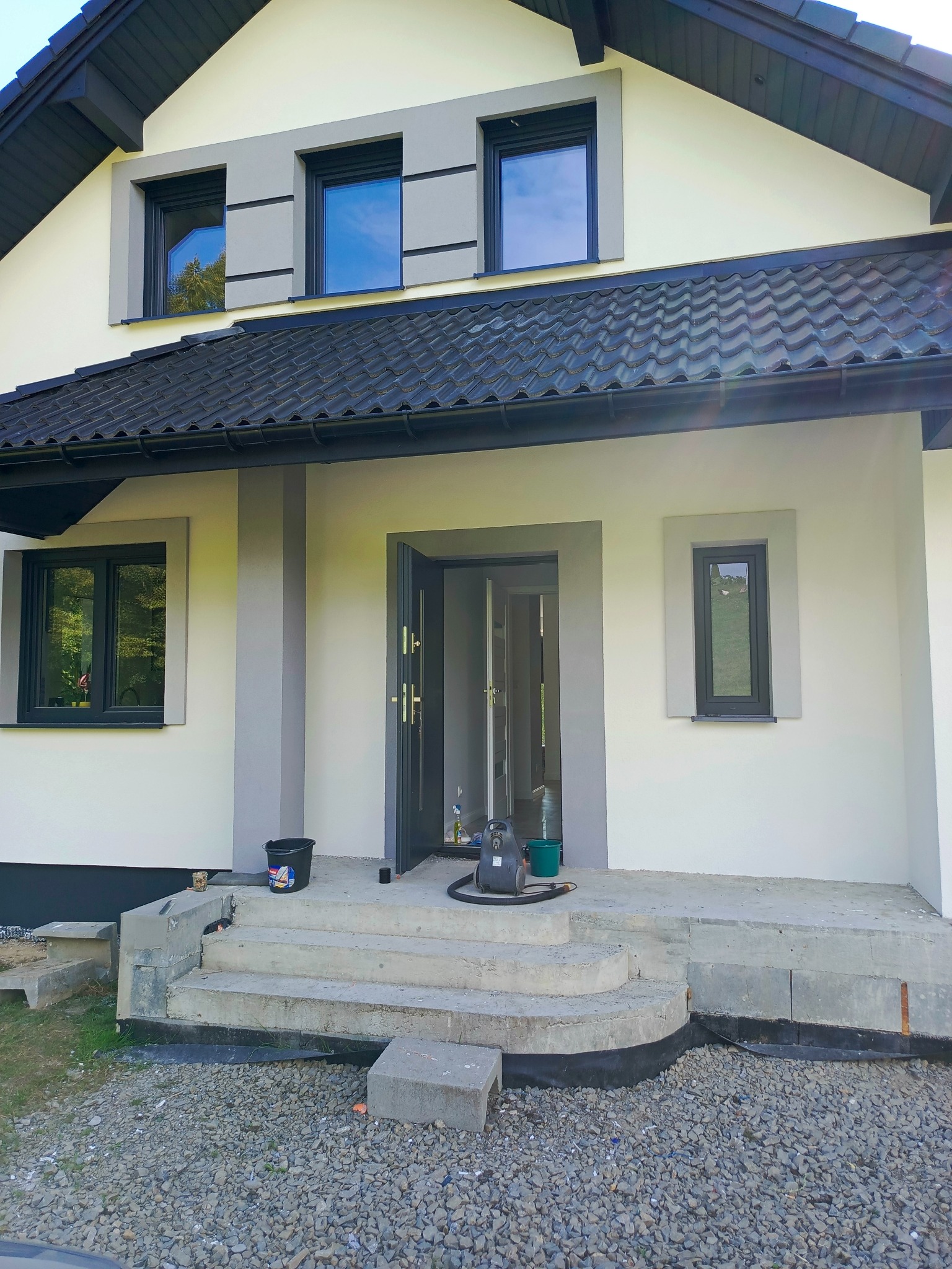Dom jednorodzinny po remoncie elewacji z szarymi obramowaniami okien i drzwi. Widoczne schody wejściowe w trakcie prac wykończeniowych.