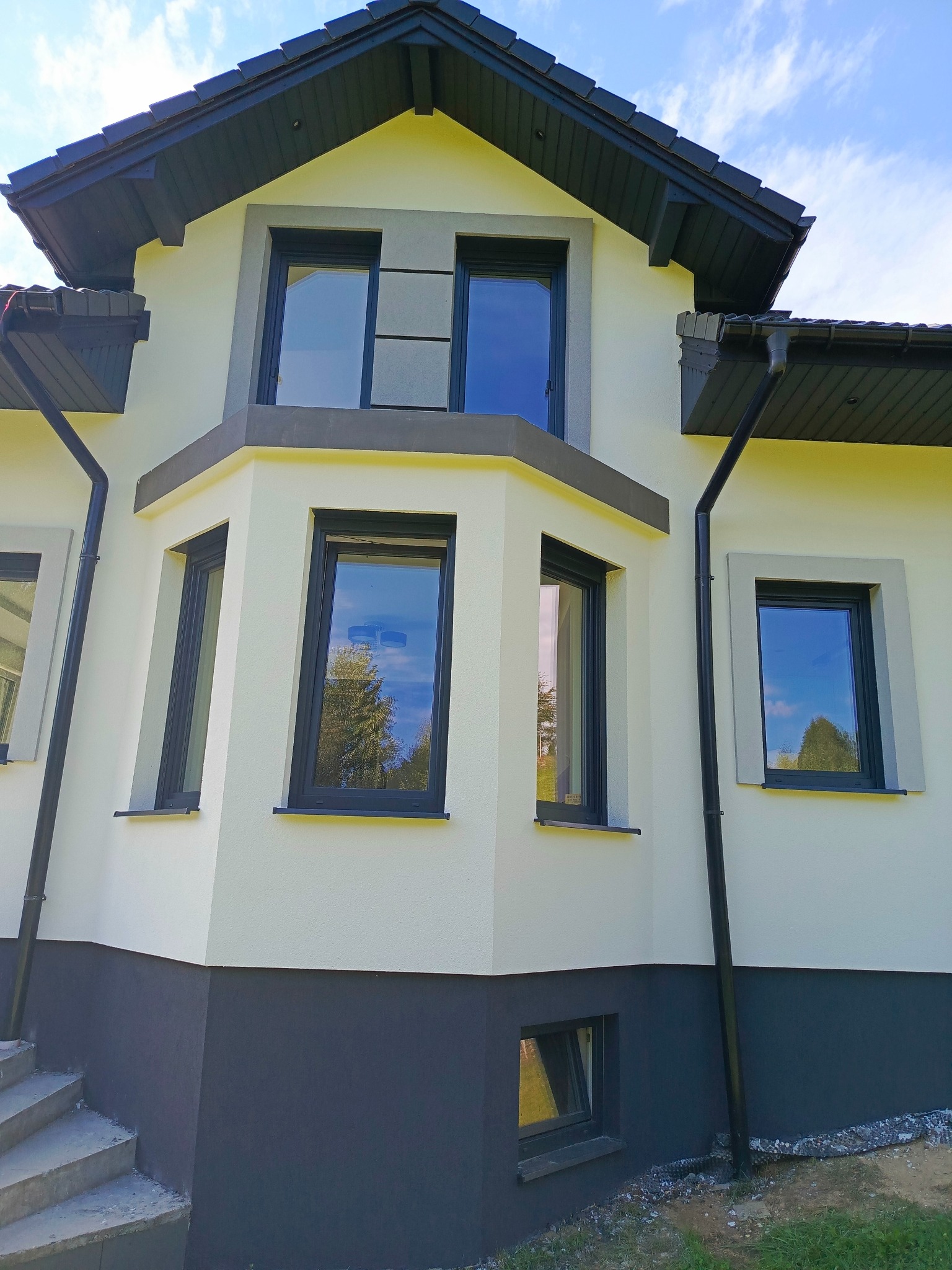 Elewacja domu jednorodzinnego po remoncie: jasna fasada z ciemną podmurówką i stolarką okienną. Widoczne rynny i fragment schodów wejściowych. Błękitne niebo odbija się w szybach.