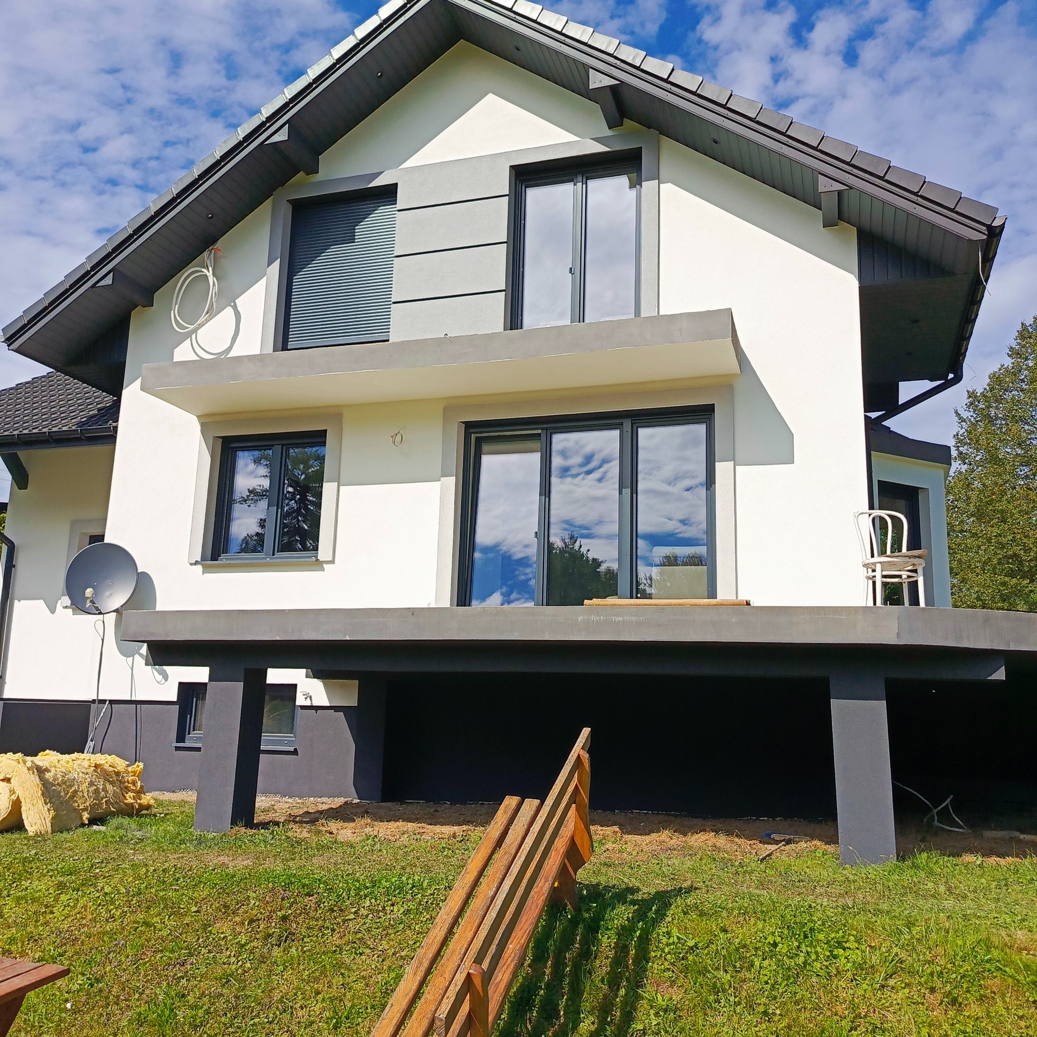 Nowoczesny dom z białoszarą elewacją i czarnymi oknami. Widoczne ocieplenie i elementy rusztowania, sugerujące prace wykończeniowe. Błękitne niebo z chmurami w tle.