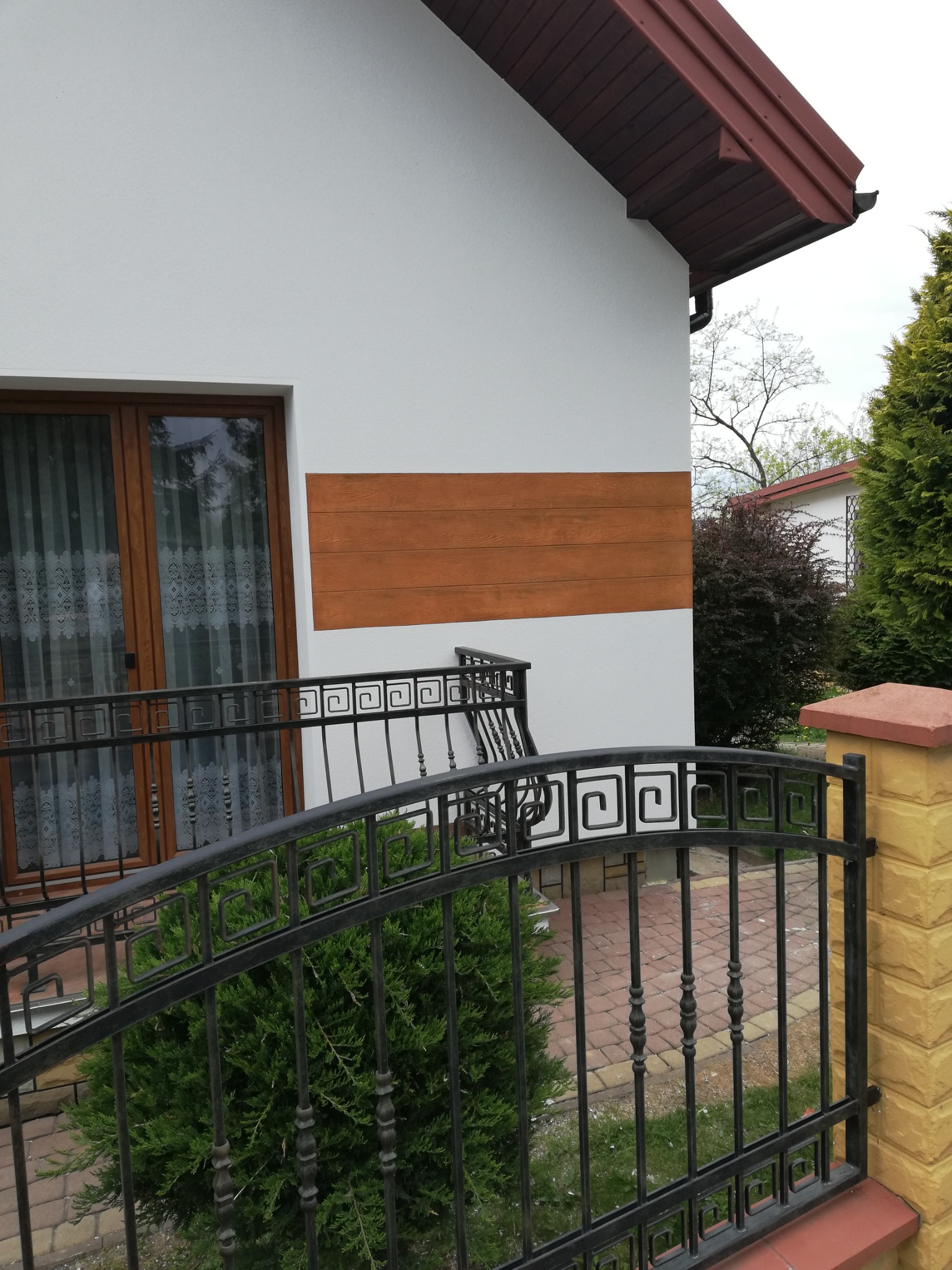Fragment domu z białą elewacją, brązowym pasem dekoracyjnym, oknami balkonowymi z firankami i czarną metalową balustradą z geometrycznym wzorem.
