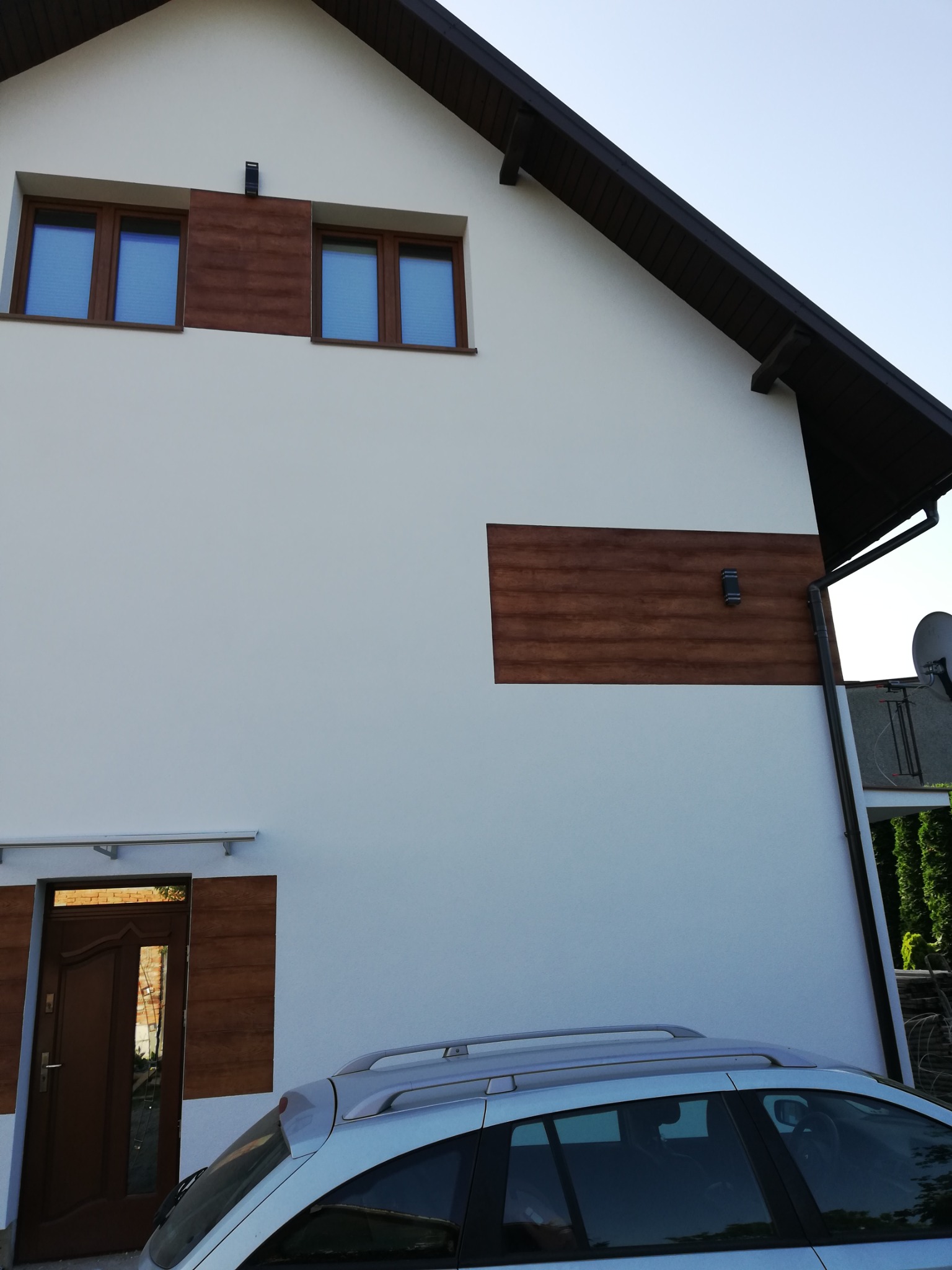 Biała elewacja domu jednorodzinnego z brązowymi akcentami drewnopodobnymi wokół okien, drzwi wejściowych i na ścianie, widoczny fragment zaparkowanego samochodu.