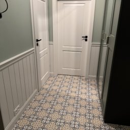 Budprint sp. z o.o. - Korytarz z wzorzystą, kremowo-szarą podłogą, białą boazerią i dwiema białymi parami drzwi z czarnymi klamkami. Ściany w kolorze jasnej zieleni.