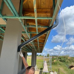 Budprint sp. z o.o. - Montaż rynny na elewacji domu jednorodzinnego. Widoczne rusztowania, drewniana konstrukcja dachu i fragment krajobrazu. Prace wykończeniowe na zewnątrz budynku.