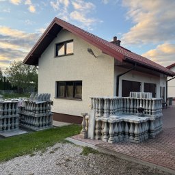 Budprint sp. z o.o. - Dom jednorodzinny z beżową elewacją i stertą betonowych balustrad na posesji. Widoczne elementy dekoracyjne i wykończeniowe. Ujęcie w słoneczny dzień.