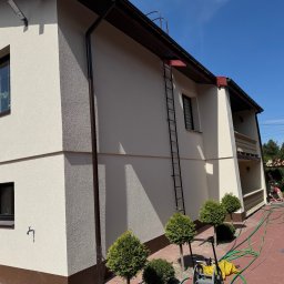 Budprint sp. z o.o. - Elewacja domu jednorodzinnego w jasnych barwach, z widoczną fakturą tynku i brązowymi akcentami. Na ścianie drabina, obok małe drzewka i wąż ogrodowy.