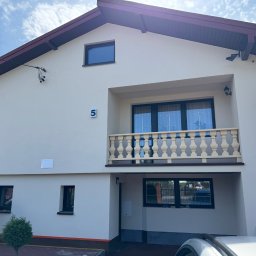 Budprint sp. z o.o. - Dwupiętrowy dom z balkonem i numerem '5' na elewacji. Dwóch mężczyzn przy oknie. Jasna elewacja, ciemne okna i dach. Częściowo widoczny samochód.