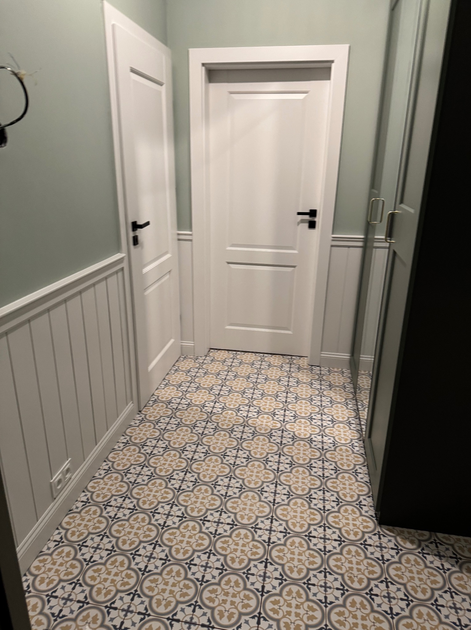 Korytarz z wzorzystą, kremowo-szarą podłogą, białą boazerią i dwiema białymi parami drzwi z czarnymi klamkami. Ściany w kolorze jasnej zieleni.