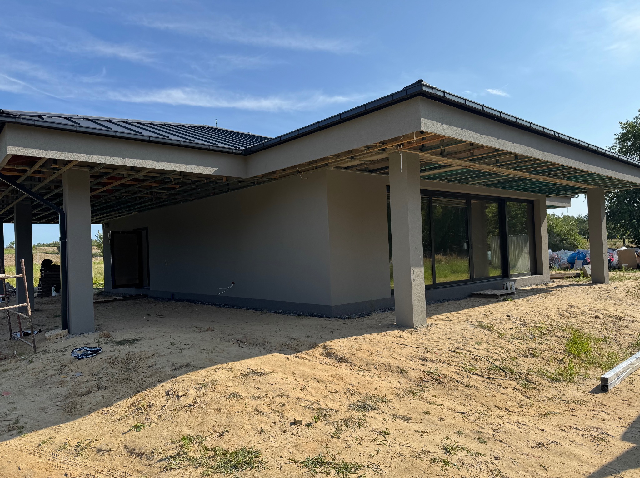 Nowoczesny dom jednorodzinny w trakcie wykończenia elewacji zewnętrznej, widoczne szare ściany, czarne okna i elementy konstrukcyjne dachu. Teren wokół w stanie surowym.