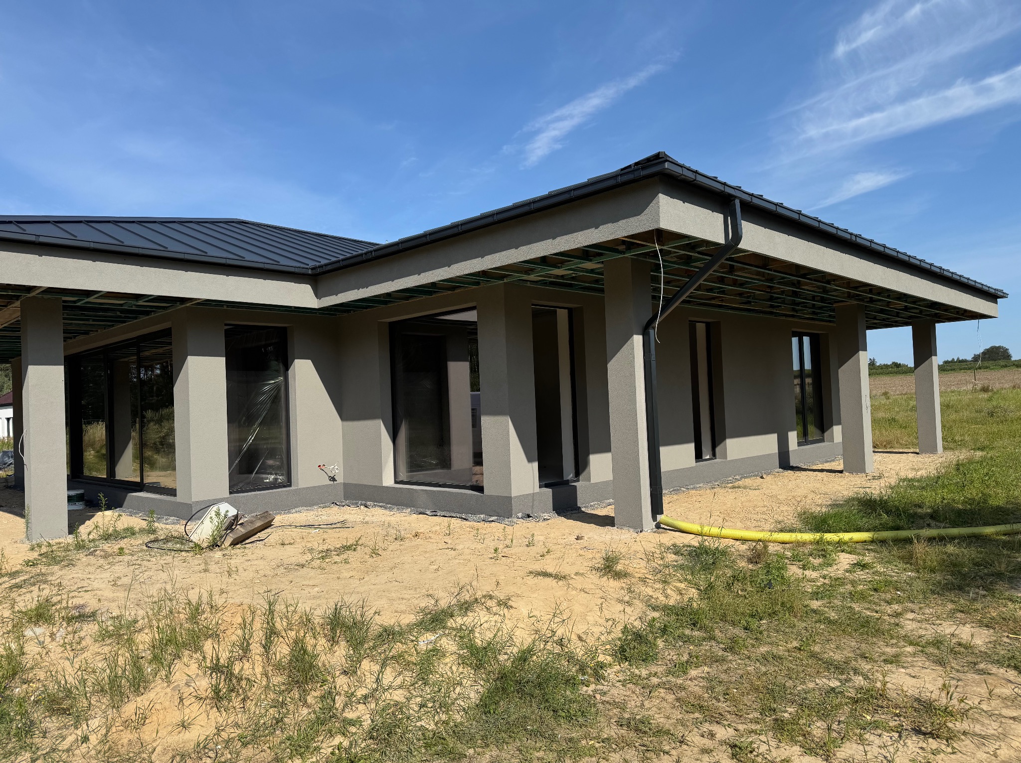 Nowoczesny dom parterowy z szarą elewacją i czarnym dachem, w trakcie prac wykończeniowych. Widoczne okna zabezpieczone folią i żółty wąż na trawniku.