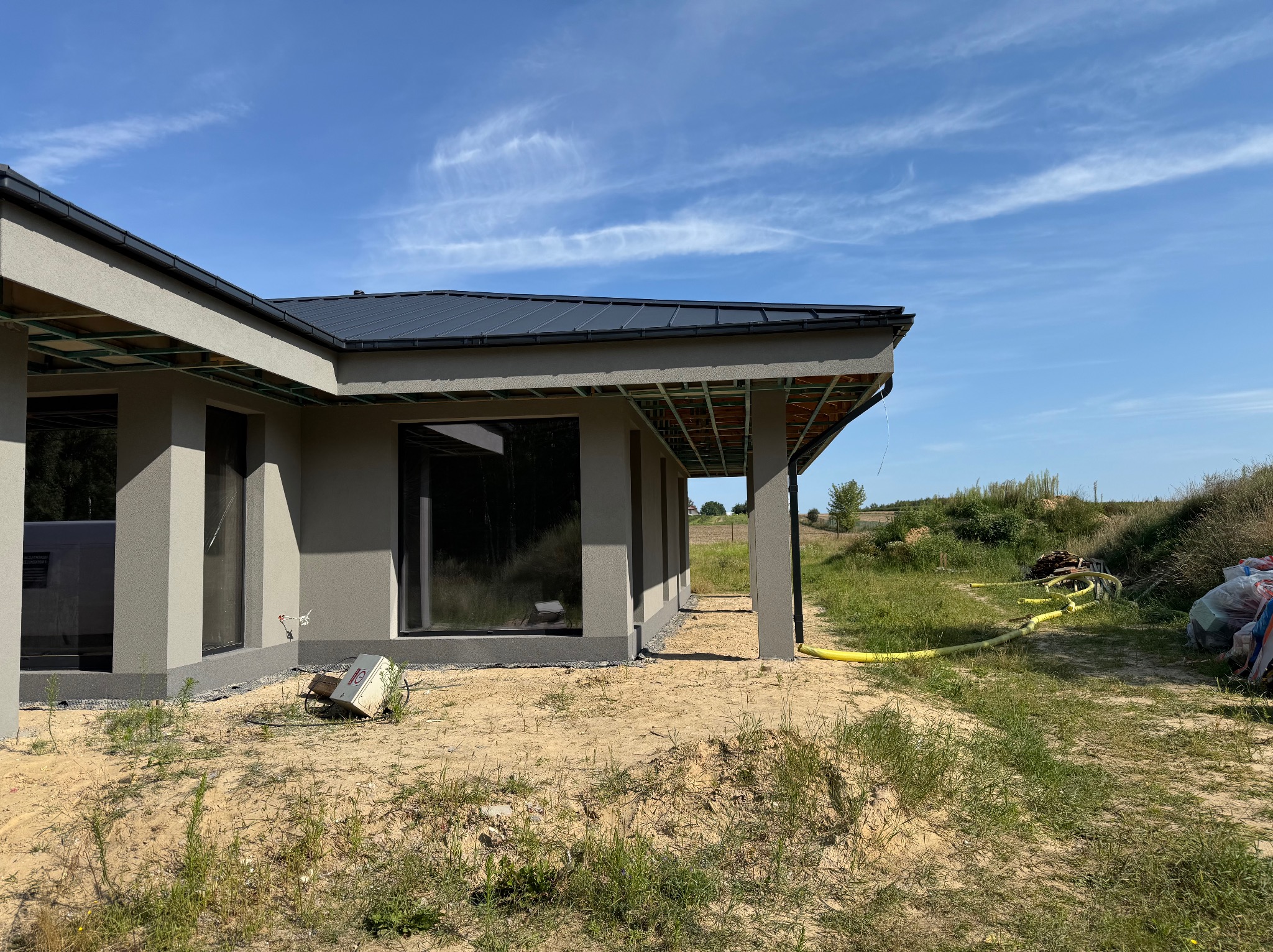 Nowoczesny dom z szarą elewacją i dużymi oknami, widok z zewnątrz w słoneczny dzień. Widoczne elementy konstrukcyjne dachu i otoczenie w trakcie prac wykończeniowych.