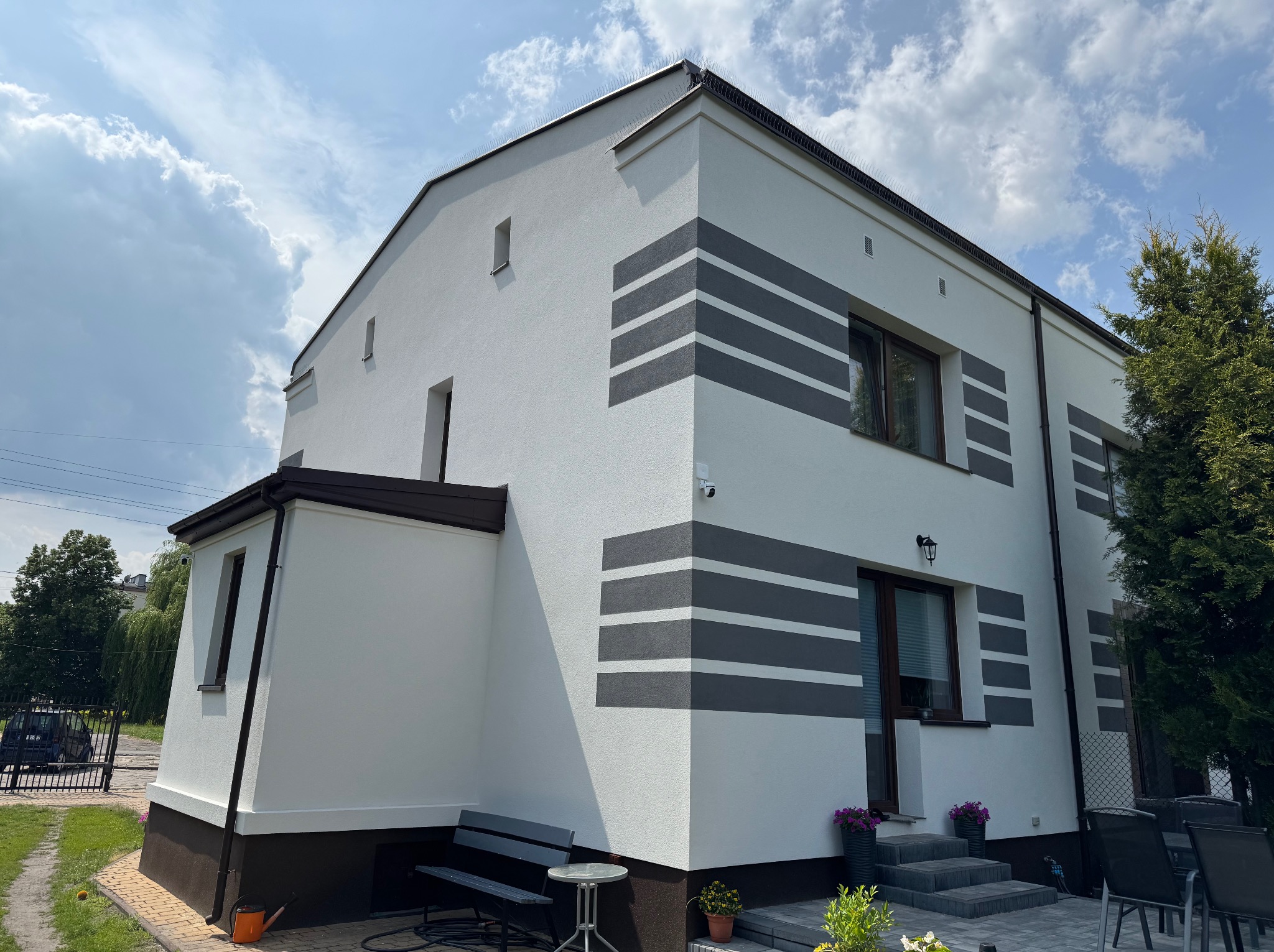 Biała elewacja domu z szarymi, poziomymi pasami dekoracyjnymi, widok z zewnątrz w słoneczny dzień. Uwagę przykuwają detale wykończenia i nowoczesny design.