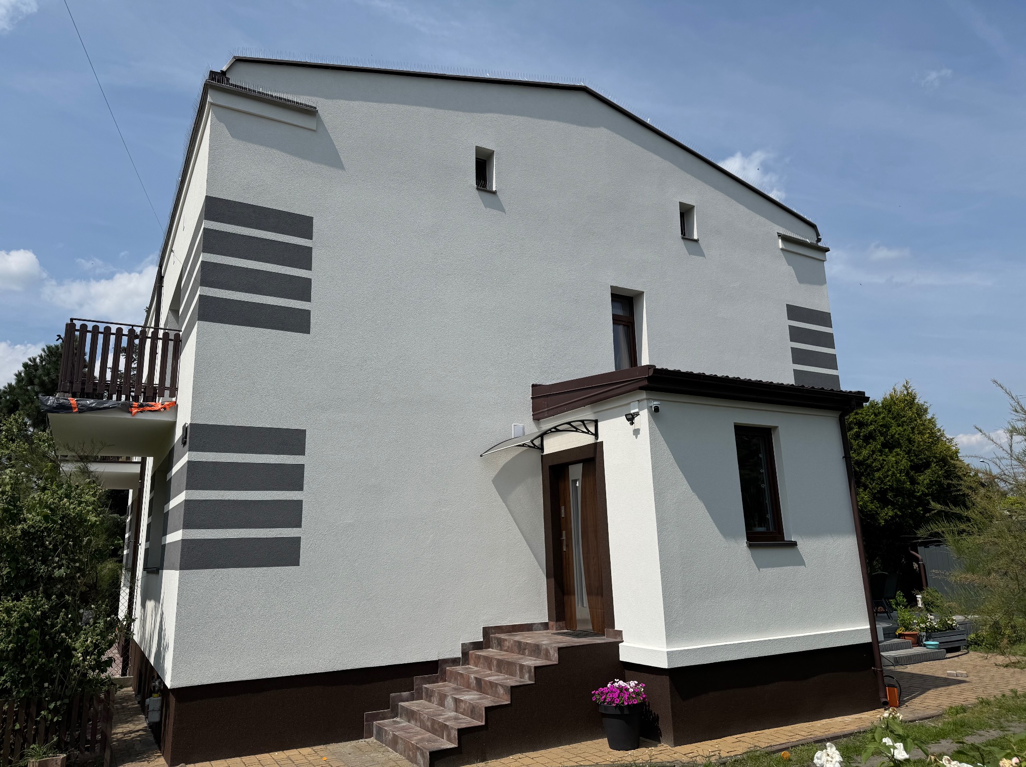 Biały dom z szarymi pasami i brązowymi akcentami, widok z dołu, podkreślający nowoczesną elewację i schody wejściowe z donicą kwiatów.