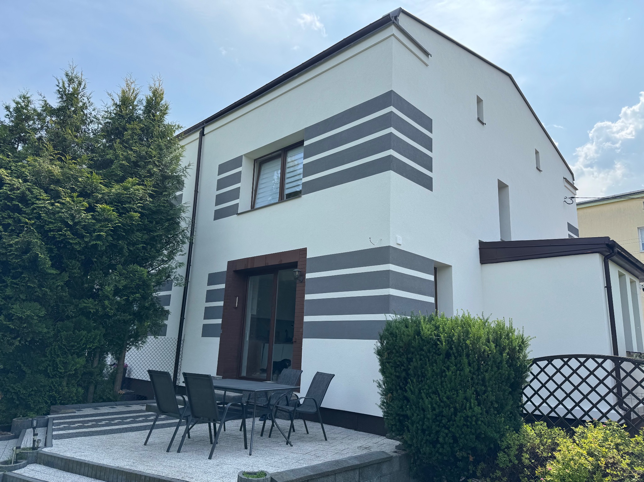 Biała elewacja domu z szarymi pasami, brązowe obramowania okien i drzwi, stół z krzesłami na tarasie, zieleń drzew i krzewów. Nowoczesny design.