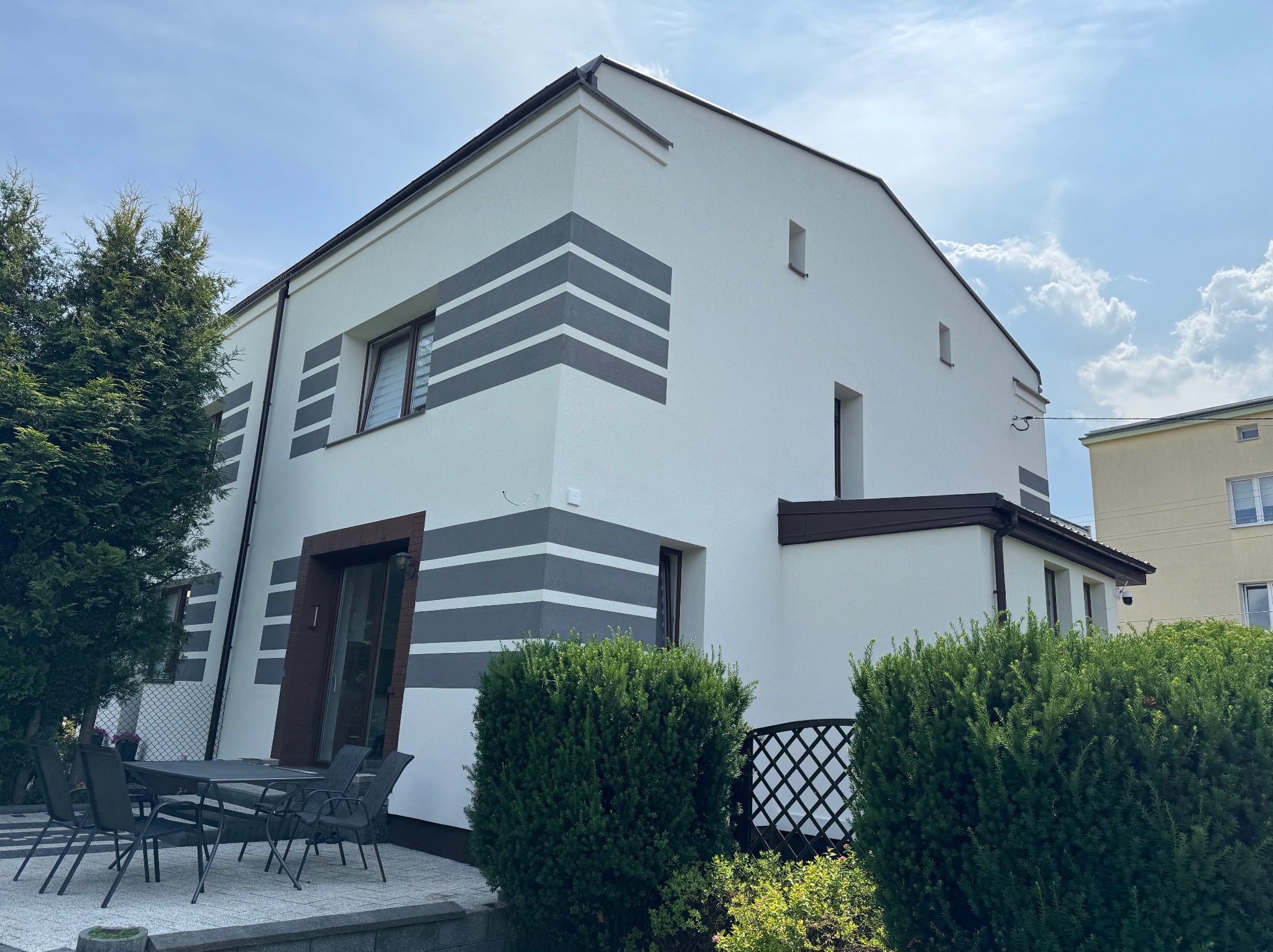 Nowoczesny dom po renowacji elewacji, z białymi ścianami i szarymi pasami dekoracyjnymi. Widoczne okna, drzwi i fragment tarasu z meblami ogrodowymi.
