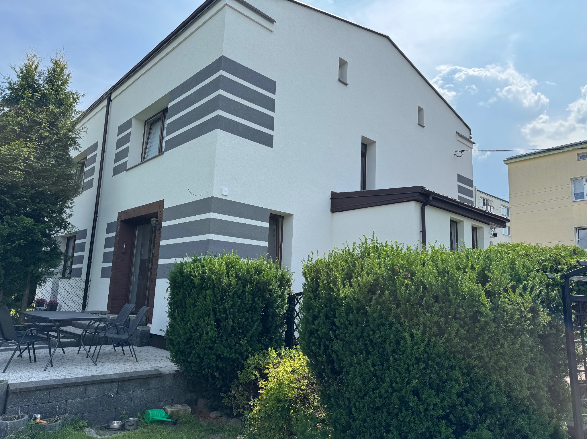 Dwupiętrowy dom z białą elewacją i szarymi pasami, widoczny z poziomu ogrodu z bujną zielenią. Stolik z krzesłami na tarasie z boku budynku.