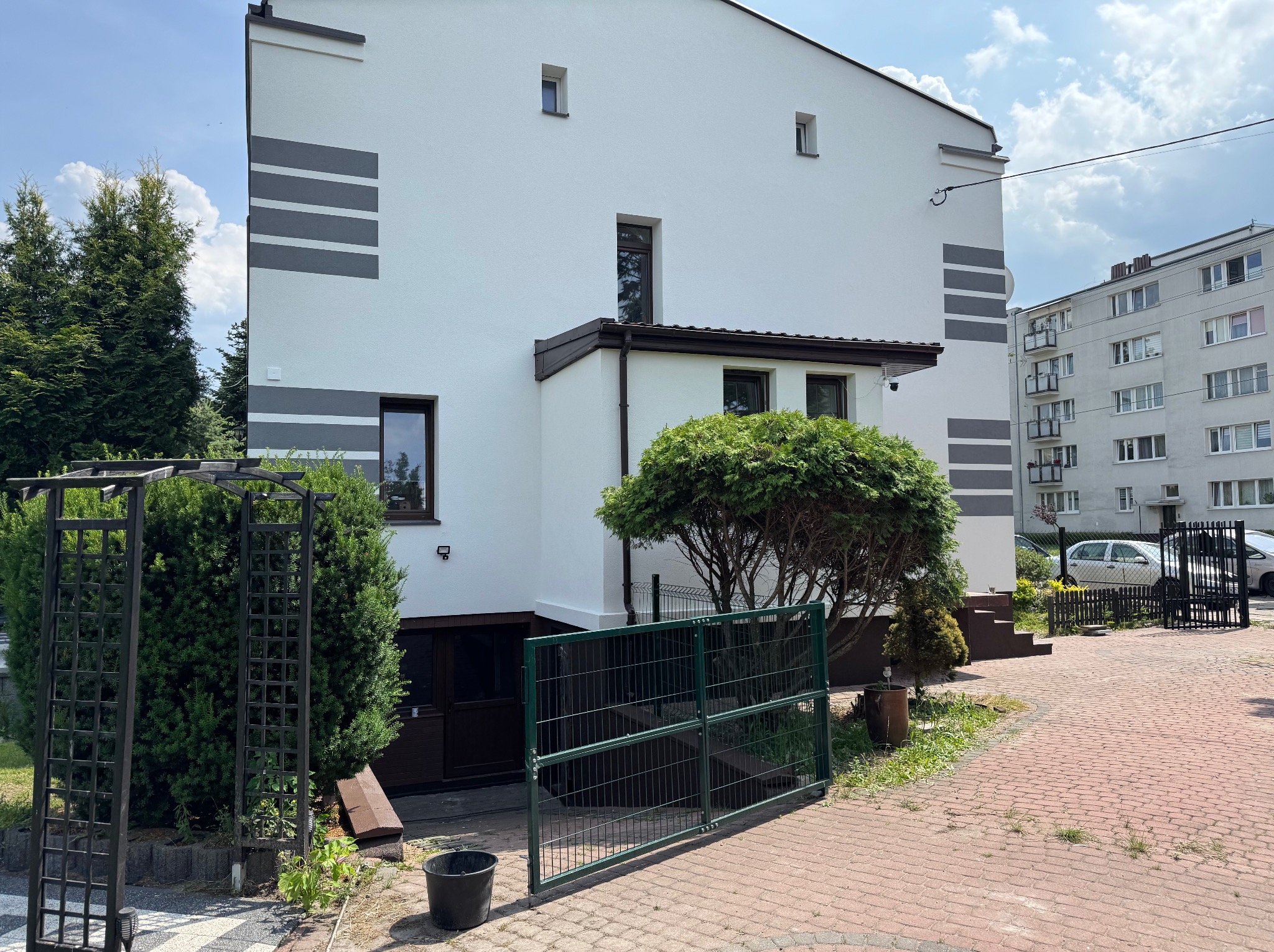 Biała elewacja domu z szarymi pasami dekoracyjnymi, widok z zewnątrz, zieleń w tle. Nowoczesny design, jasne kolory, brama wjazdowa.