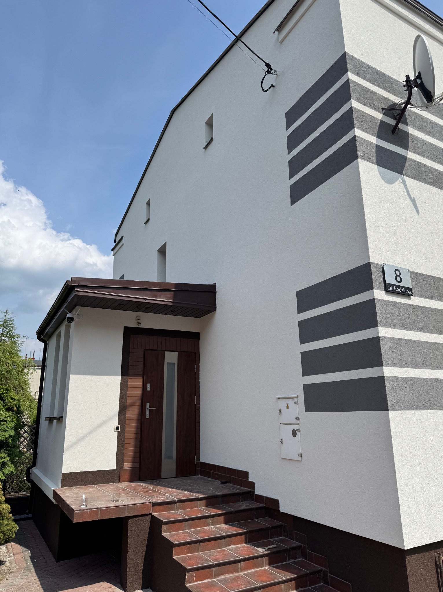 Biała elewacja domu z szarymi pasami, brązowe drzwi wejściowe i schody z płytek. Widoczny numer domu i antena satelitarna. Słoneczny dzień.