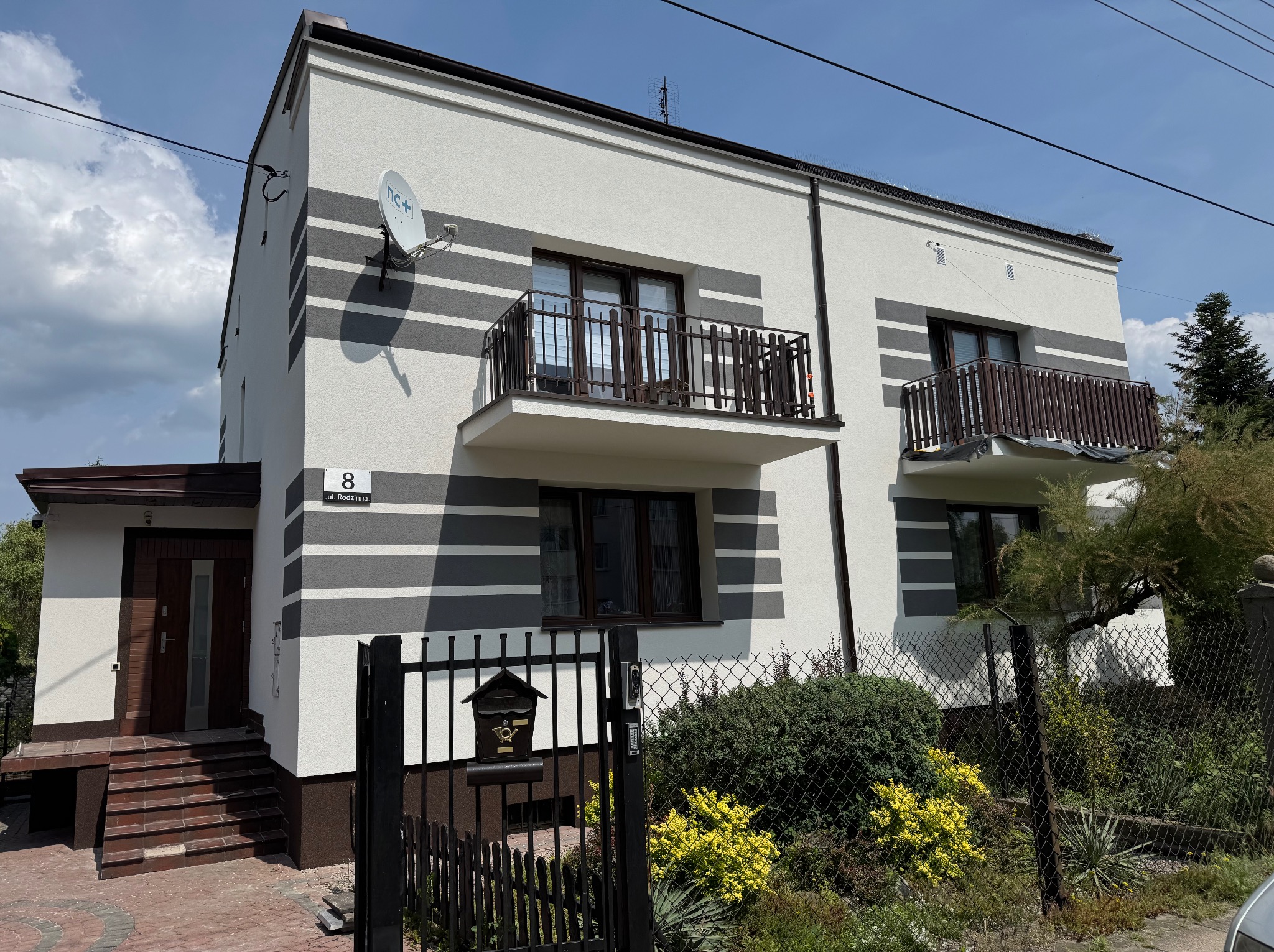Dwupiętrowy dom z balkonami i elewacją w poziome pasy szarości i bieli, widoczny zza ogrodzenia z furtką i skrzynką na listy. Na ścianie numer domu i antena satelitarna.
