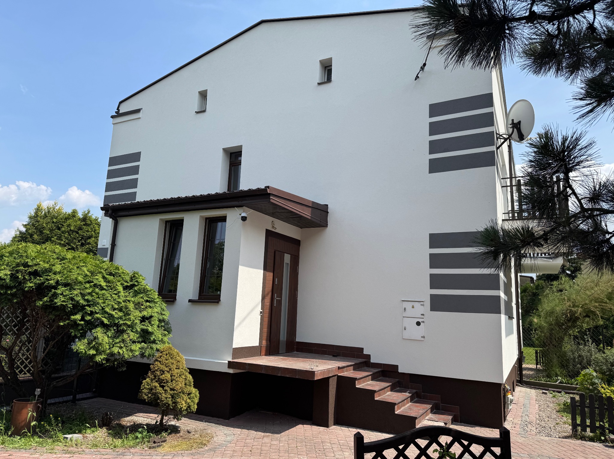 Biały dom z szarymi pasami i brązowymi elementami wykończeniowymi, widok z zewnątrz w słoneczny dzień. Widoczna antena satelitarna i zadbana zieleń.