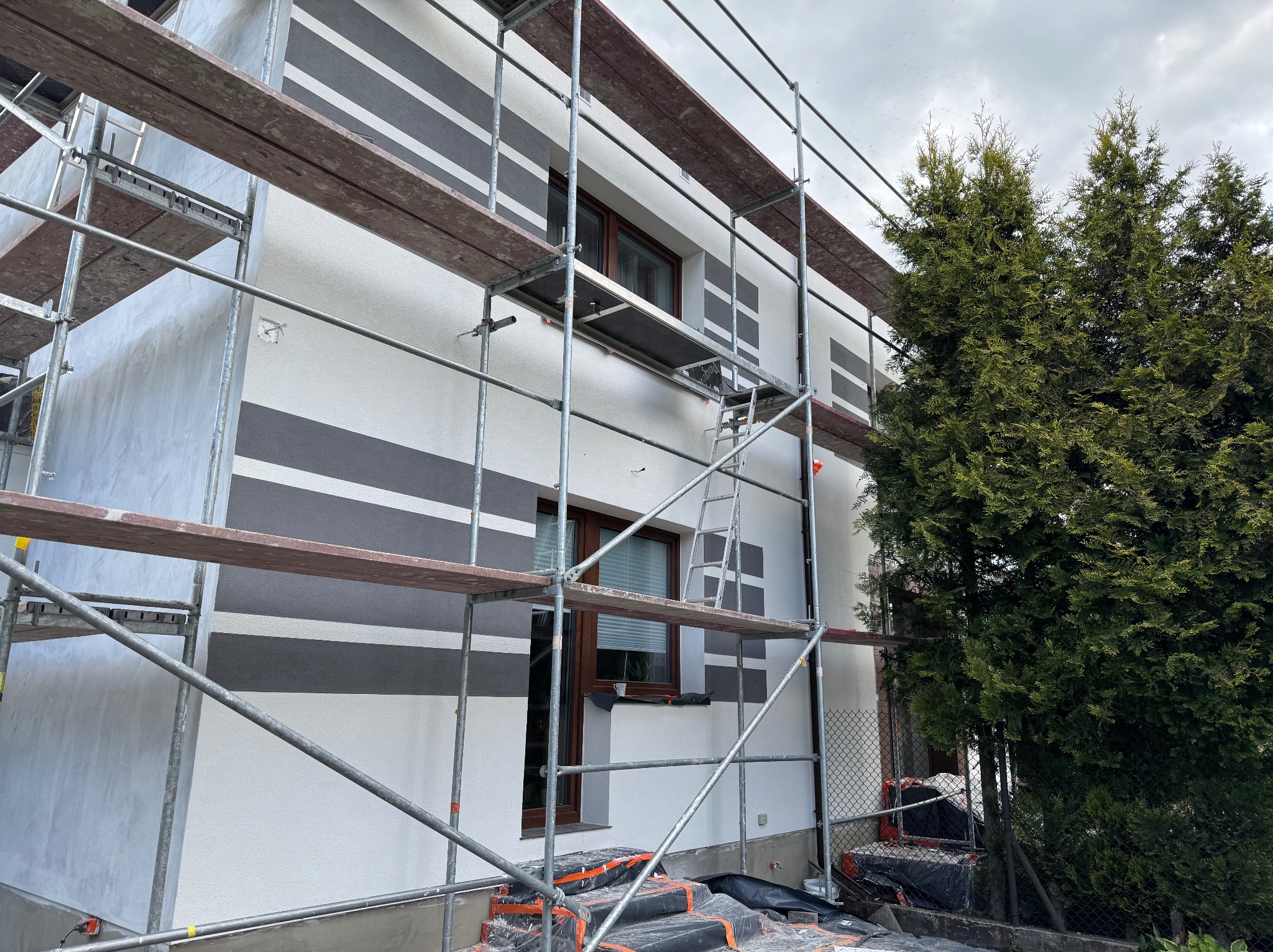 Elewacja domu w trakcie prac: biała ściana z szarymi pasami, rusztowanie, okna z brązowymi ramami. Widok z dołu, częściowo zasłonięty przez drzewo.