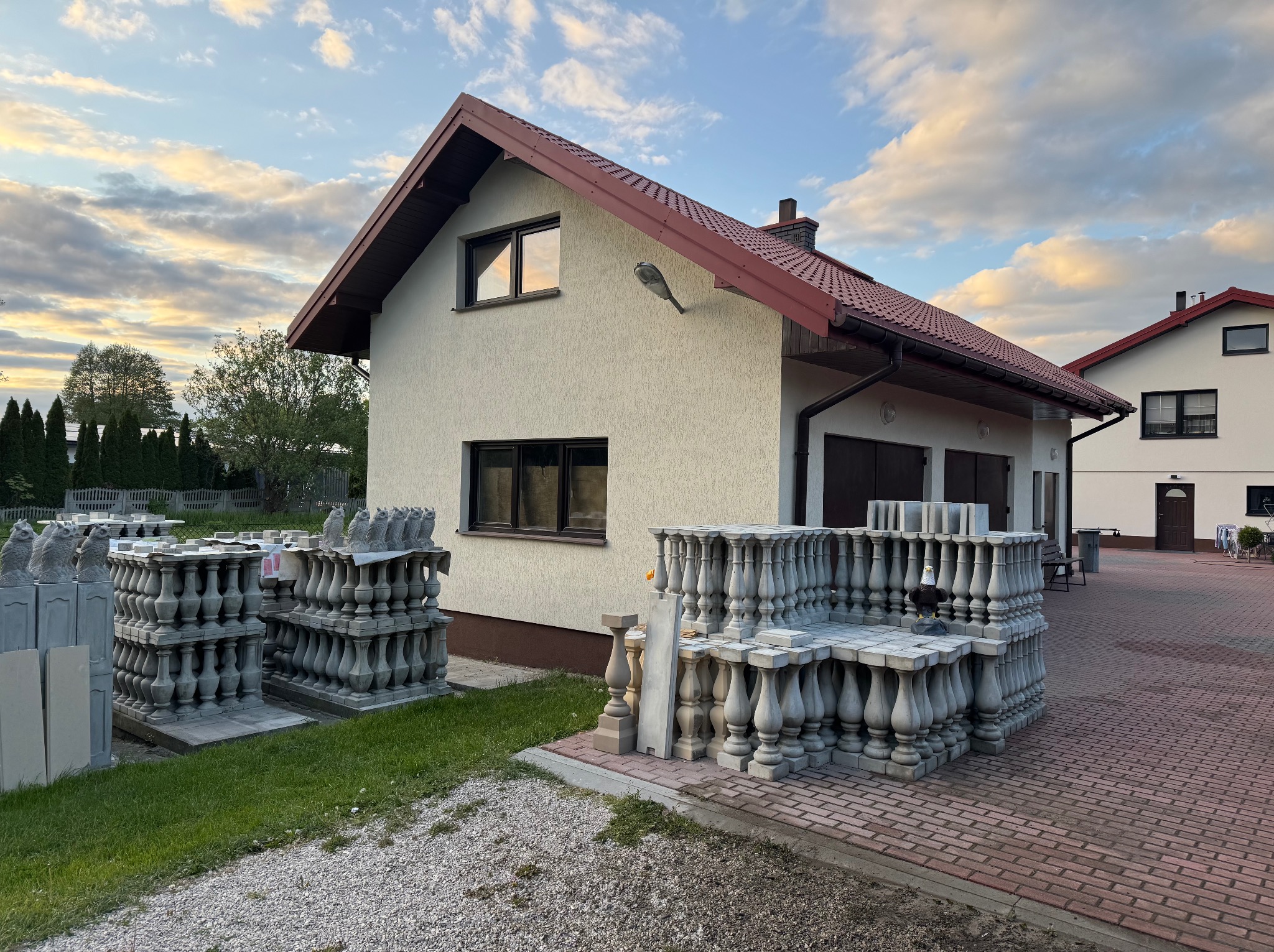 Dom jednorodzinny z beżową elewacją i stertą betonowych balustrad na posesji. Widoczne elementy dekoracyjne i wykończeniowe. Ujęcie w słoneczny dzień.