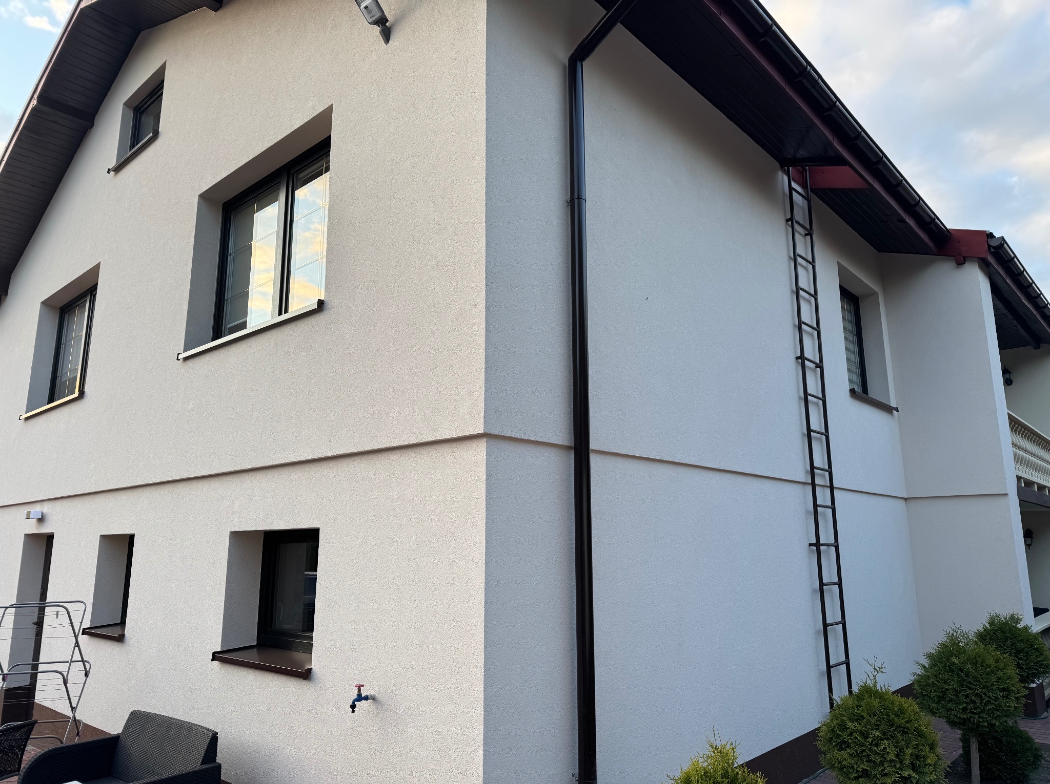 Dwukondygnacyjny dom z jasną elewacją o fakturze baranka, czarnymi oknami i rynnami. Widoczna drabina na ścianie, krzewy i fragment mebli ogrodowych.
