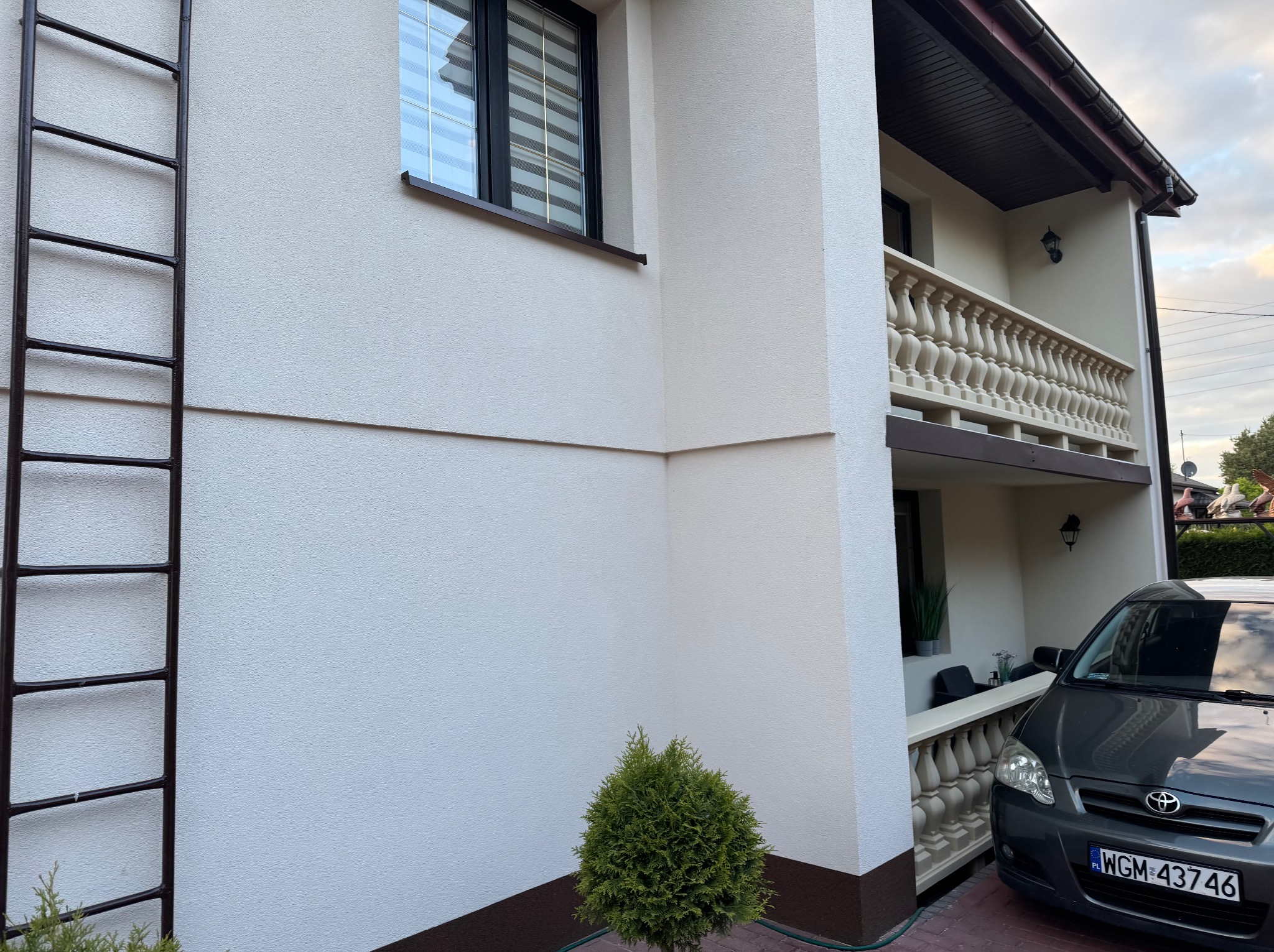 Jasna elewacja domu z balkonem i brązowymi akcentami. Widoczna drabina oparta o ścianę, mała tuja i zaparkowany samochód. Ujęcie z dołu, w tle błękitne niebo.