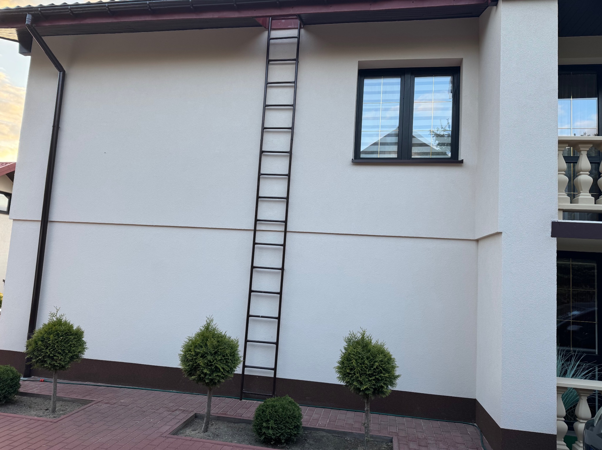 Biała elewacja domu z brązowymi akcentami, drabina oparta o ścianę, okno z roletami i trzy małe drzewka na tle czerwonej kostki brukowej.