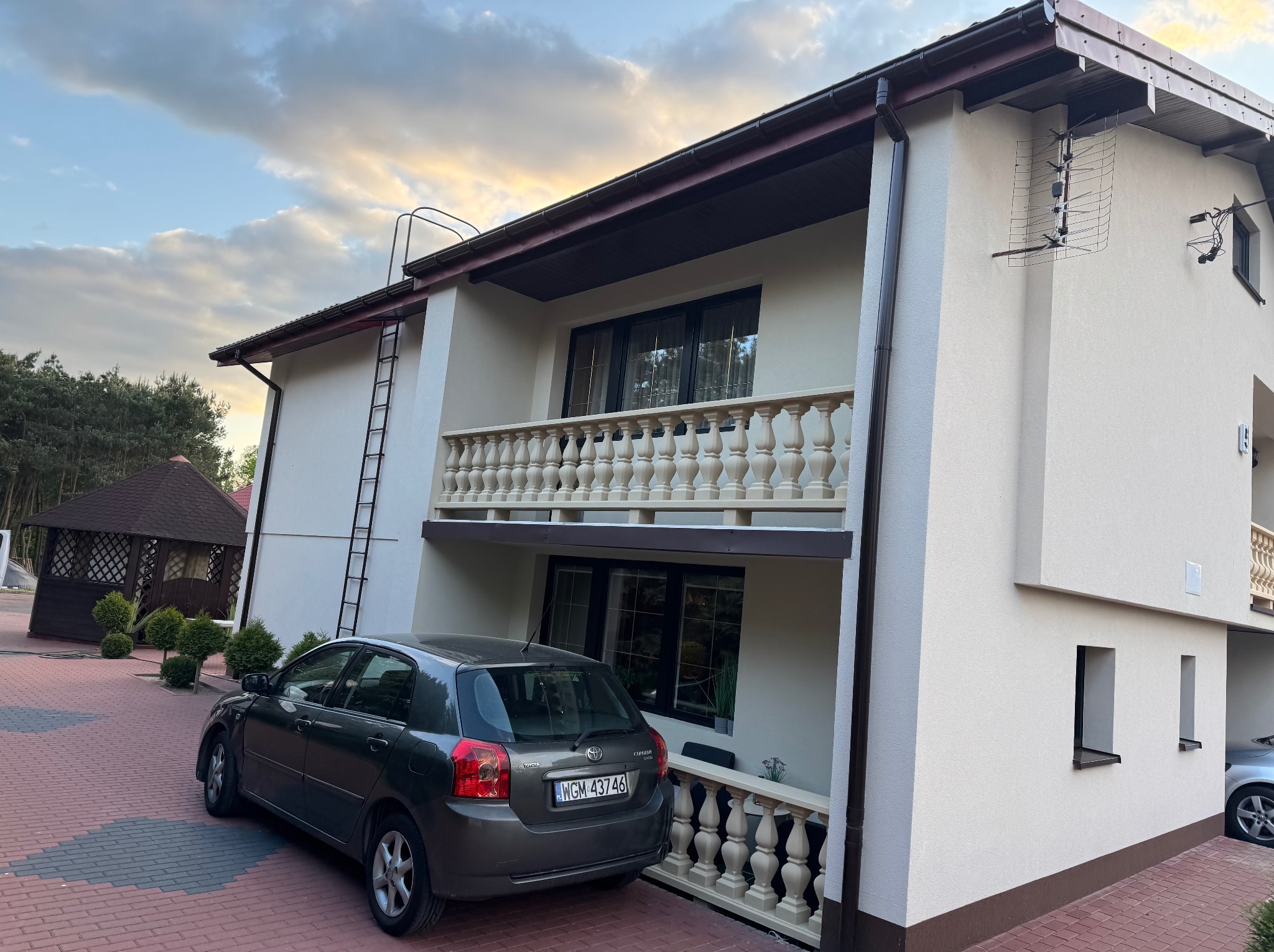 Dwupiętrowy dom z balkonami i balustradami, kremowa elewacja, brązowe okna i dach. Na ścianie antena, obok zaparkowany samochód. Widoczna altana i drzewa w tle.