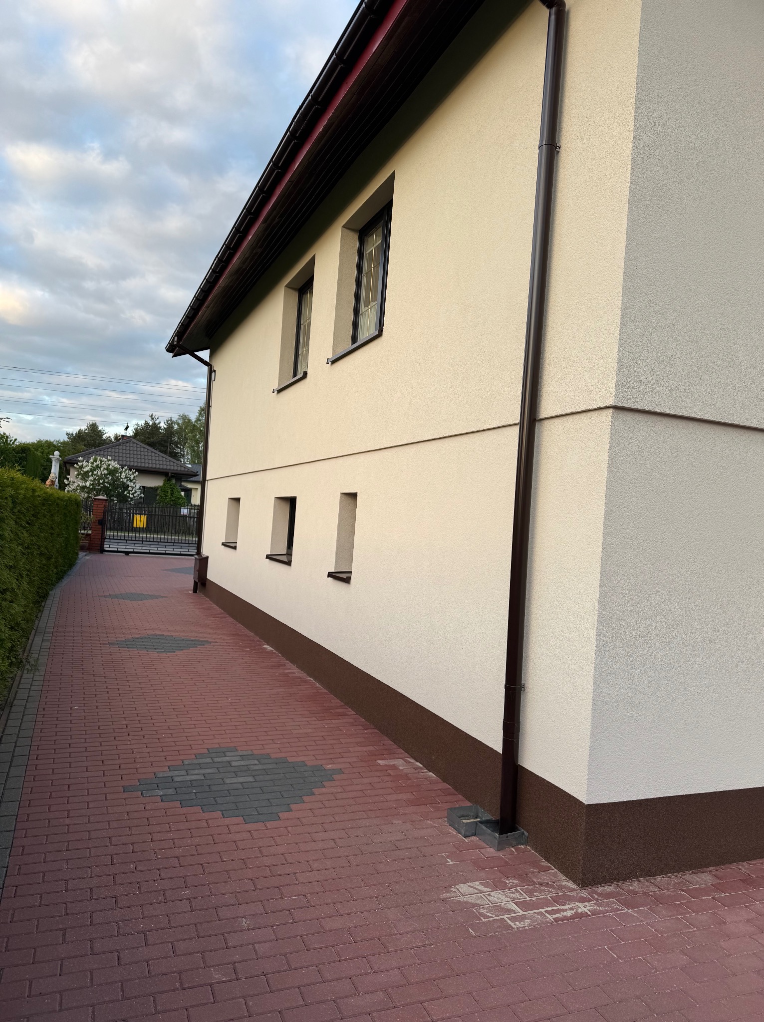 Elewacja domu jednorodzinnego w jasnym odcieniu z brązowymi detalami, widoczna z boku, z rynną i fragmentem chodnika z kostki brukowej.