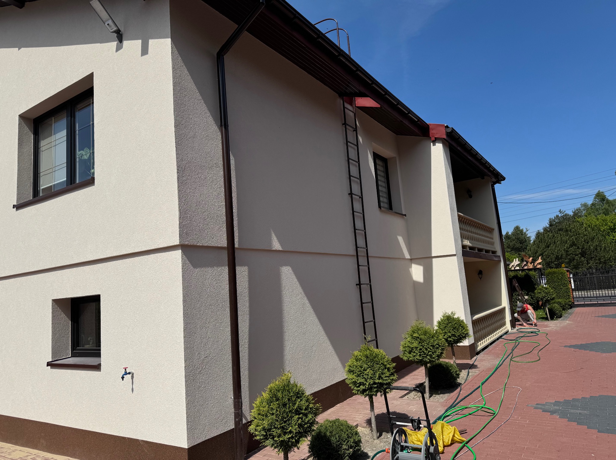 Elewacja domu jednorodzinnego w jasnych barwach, z widoczną fakturą tynku i brązowymi akcentami. Na ścianie drabina, obok małe drzewka i wąż ogrodowy.