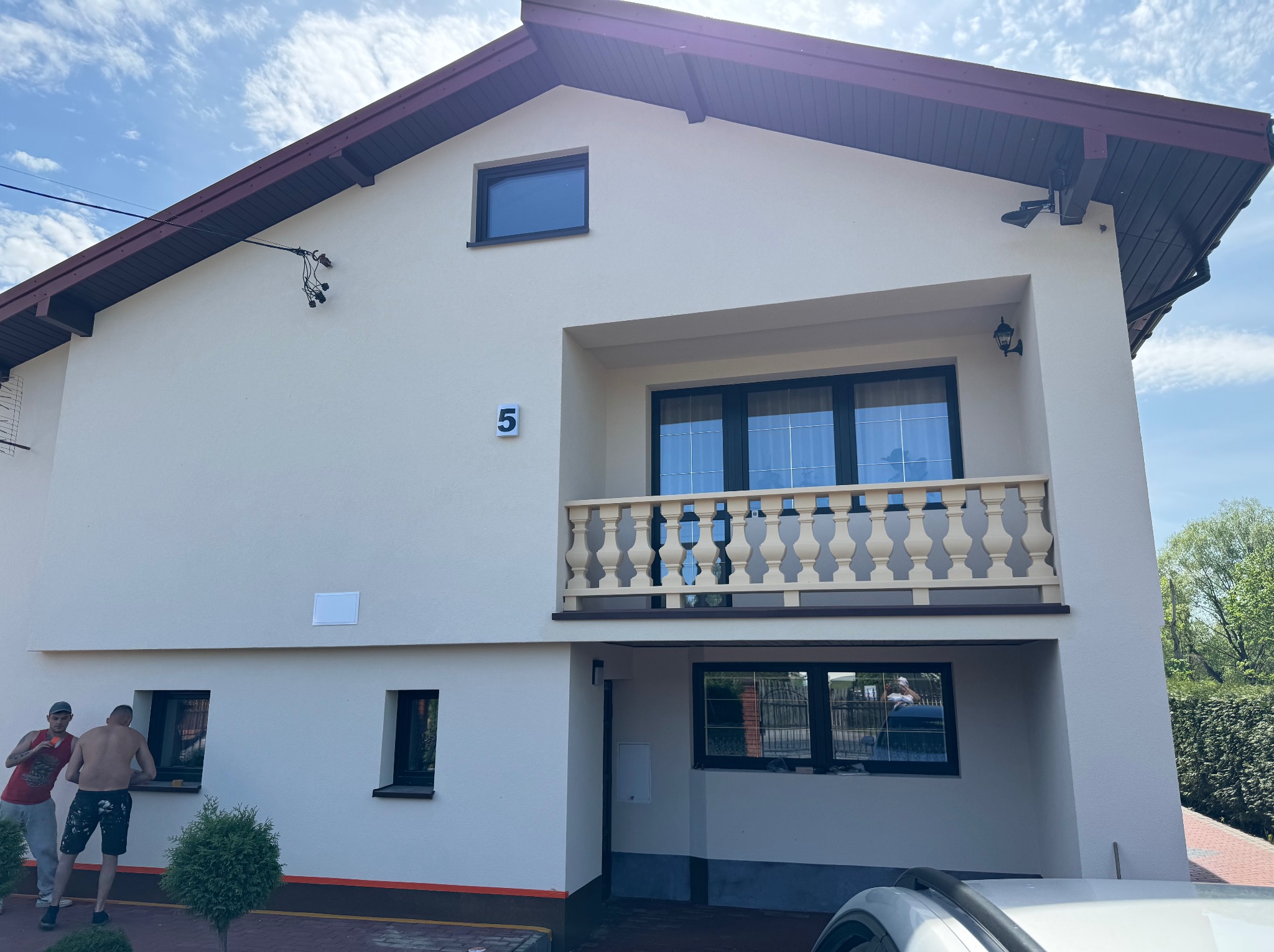 Dwupiętrowy dom z balkonem i numerem '5' na elewacji. Dwóch mężczyzn przy oknie. Jasna elewacja, ciemne okna i dach. Częściowo widoczny samochód.