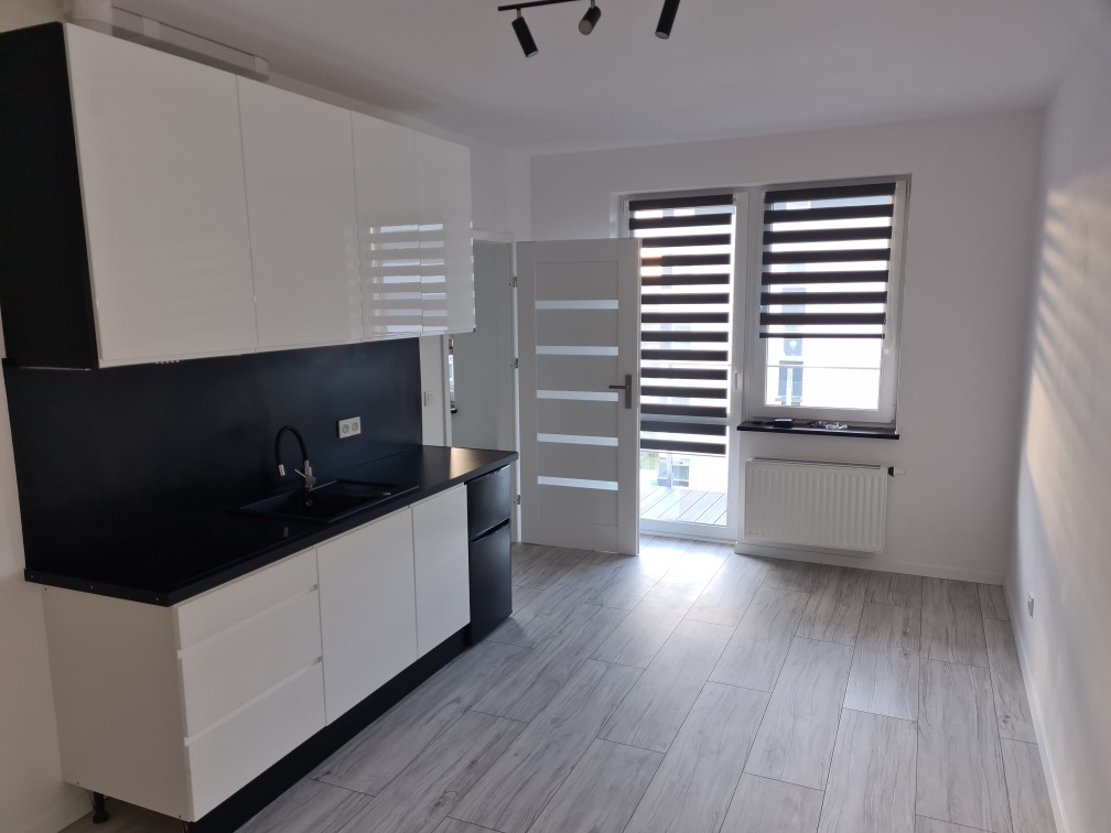 Nowoczesna kuchnia z białymi szafkami i czarnym blatem, widok na balkon przez okno z żaluzjami dzień-noc, szare panele podłogowe.