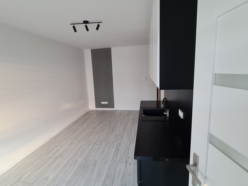 Wnętrze mieszkania z nową, czarną zabudową kuchenną, jasnymi panelami podłogowymi i nowoczesnym oświetleniem sufitowym.