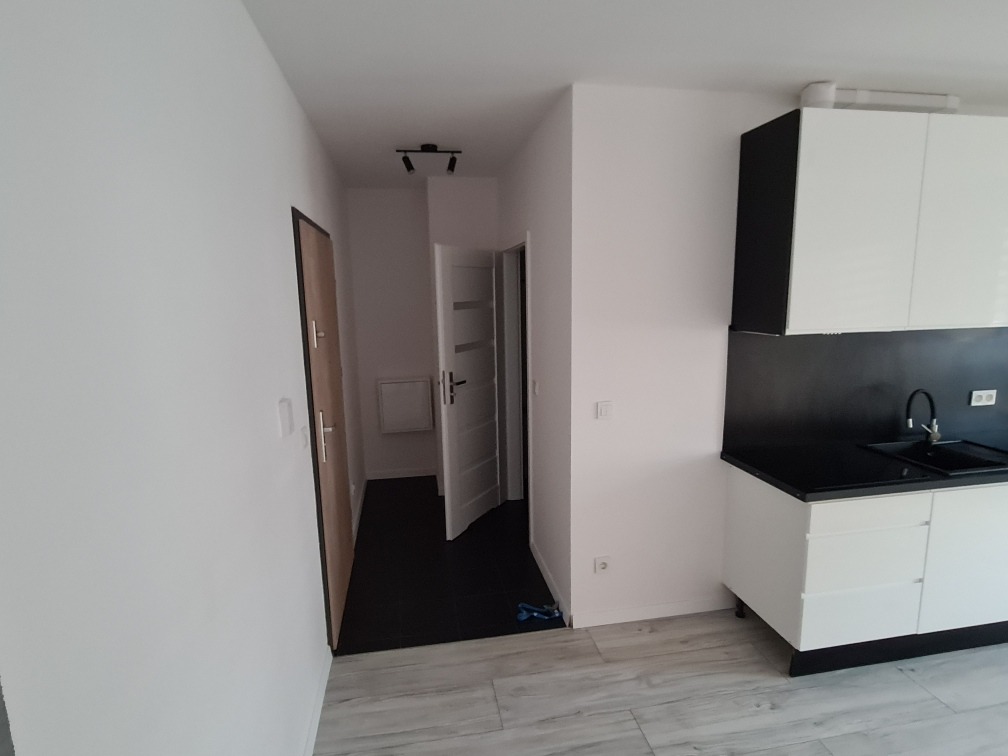 Wnętrze mieszkania po remoncie: widok na korytarz z otwartymi drzwiami i minimalistyczną zabudowę kuchenną z białymi szafkami i czarnym blatem.