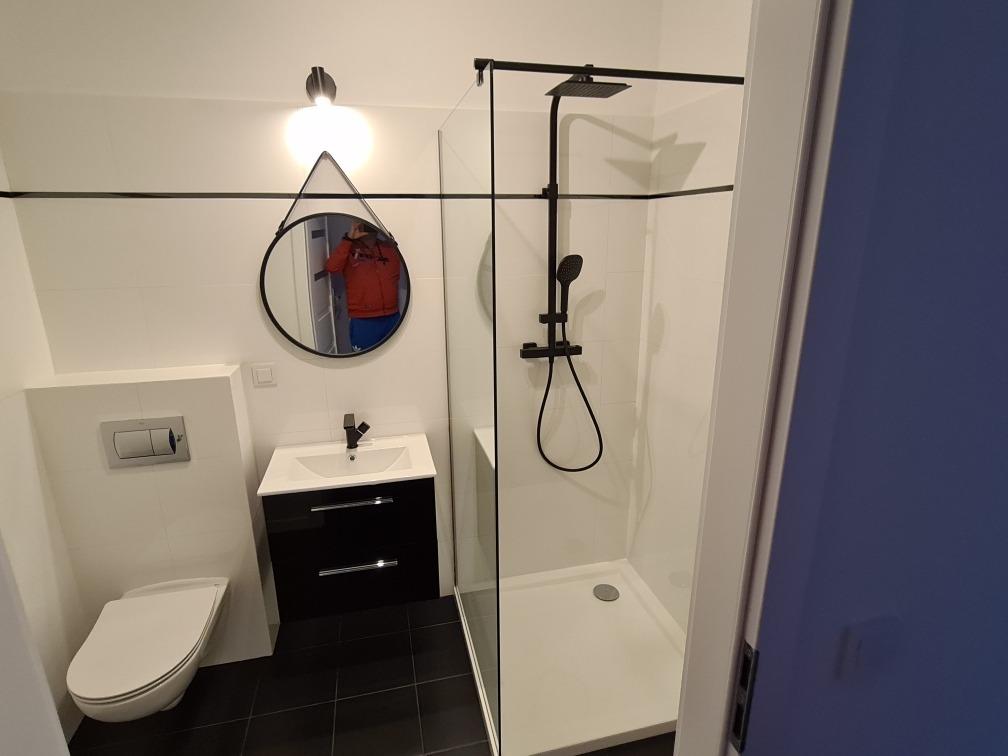 Nowoczesna łazienka z czarnymi akcentami: okrągłe lustro z trójkątnym uchwytem, czarna armatura, kabina prysznicowa typu walk-in i wisząca toaleta.