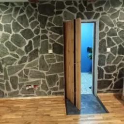 Wnętrze pokoju z kamienną ścianą z nieregularnych płyt, drewnianą podłogą parkietową i częściowo otwartymi drewnianymi drzwiami prowadzącymi do pomieszczenia z niebieską ścianą i rośliną doniczkową...