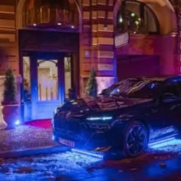 Elegancki czarny samochód z podświetleniem LED zaparkowany przed wejściem do hotelu z czerwonym dywanem i dekoracyjnymi roślinami w donicach, nocne ujęcie.