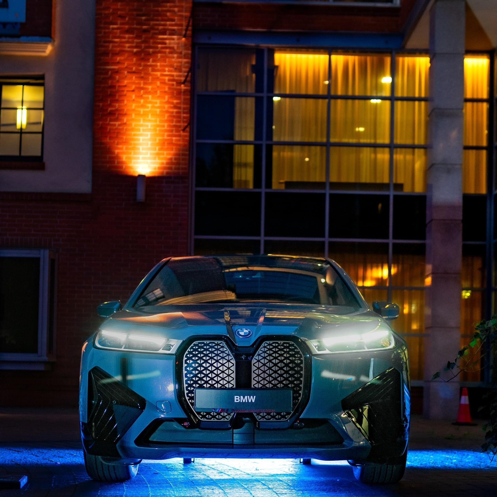 Nowoczesny, elektryczny samochód BMW iX prezentowany nocą przed budynkiem z cegły i dużymi oknami, z niebieskim oświetleniem podwozia.