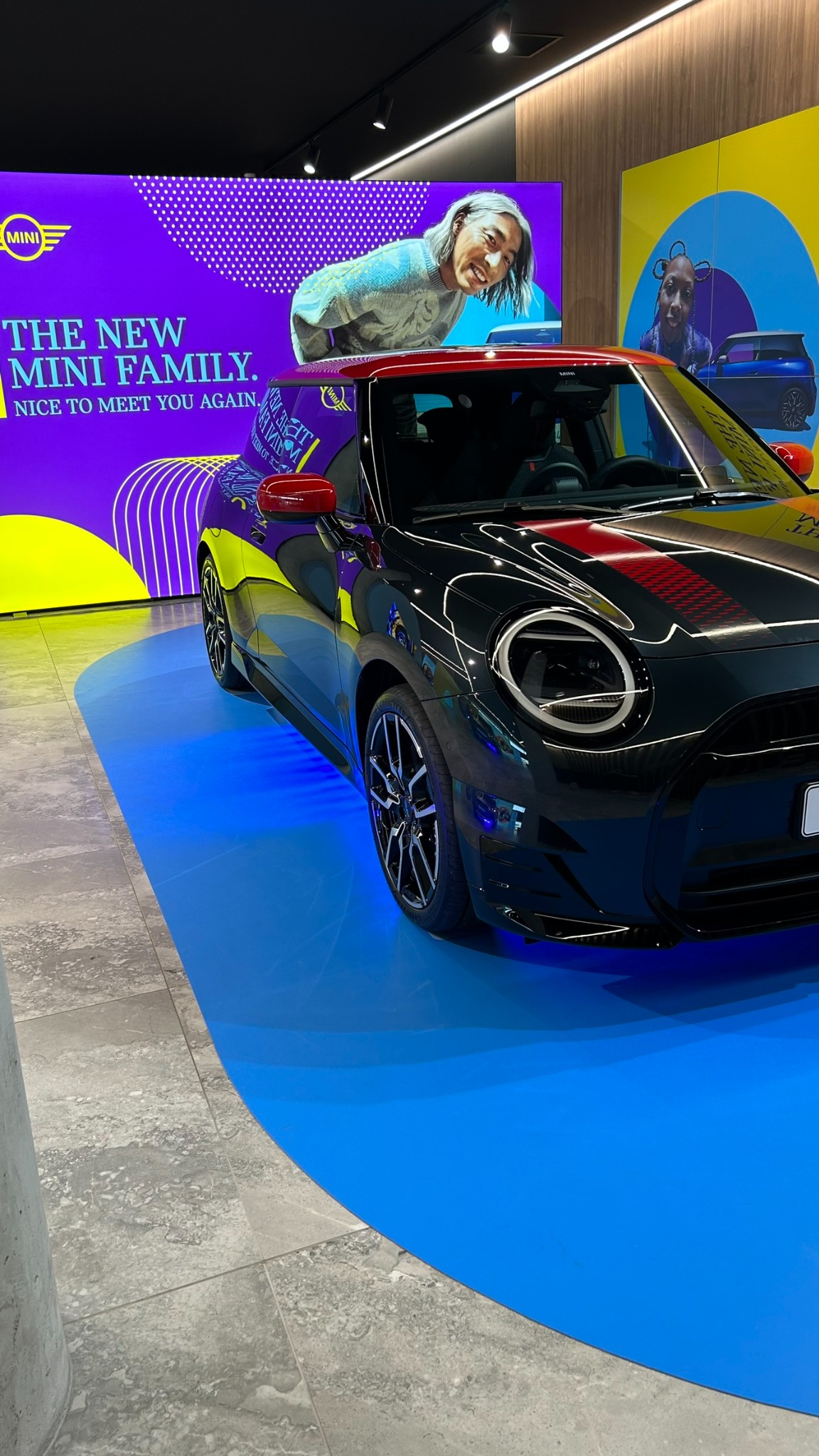 Czarne Mini Cooper z czerwonym dachem i pasem na masce, prezentowane na niebieskiej platformie w salonie z kolorowymi plakatami reklamowymi.