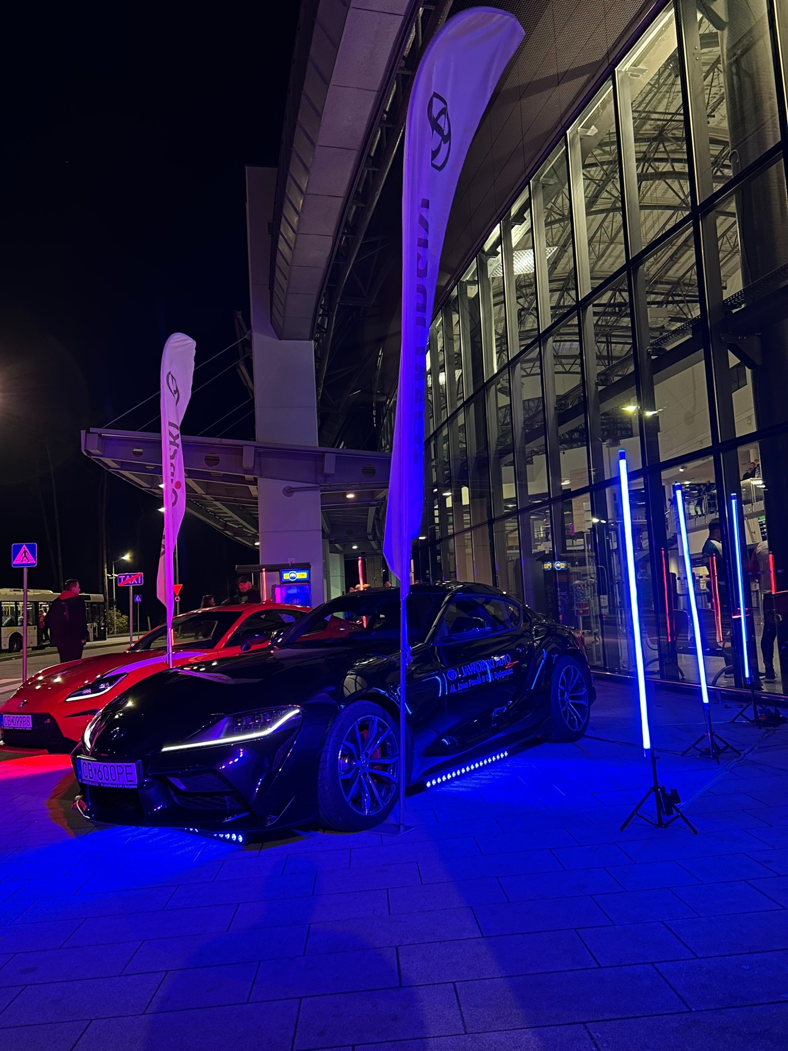 Czarny i czerwony samochód sportowy Toyota Supra oświetlone niebieskimi lampami LED przed nowoczesnym budynkiem z przeszkloną fasadą podczas nocnej imprezy.