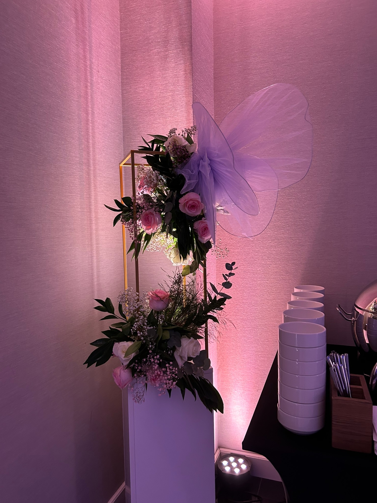 Dekoracja kwiatowa z różowych i białych róż, umieszczona na złotym stelażu, obok stosu białych talerzy i podgrzewacza, oświetlona fioletowym światłem.